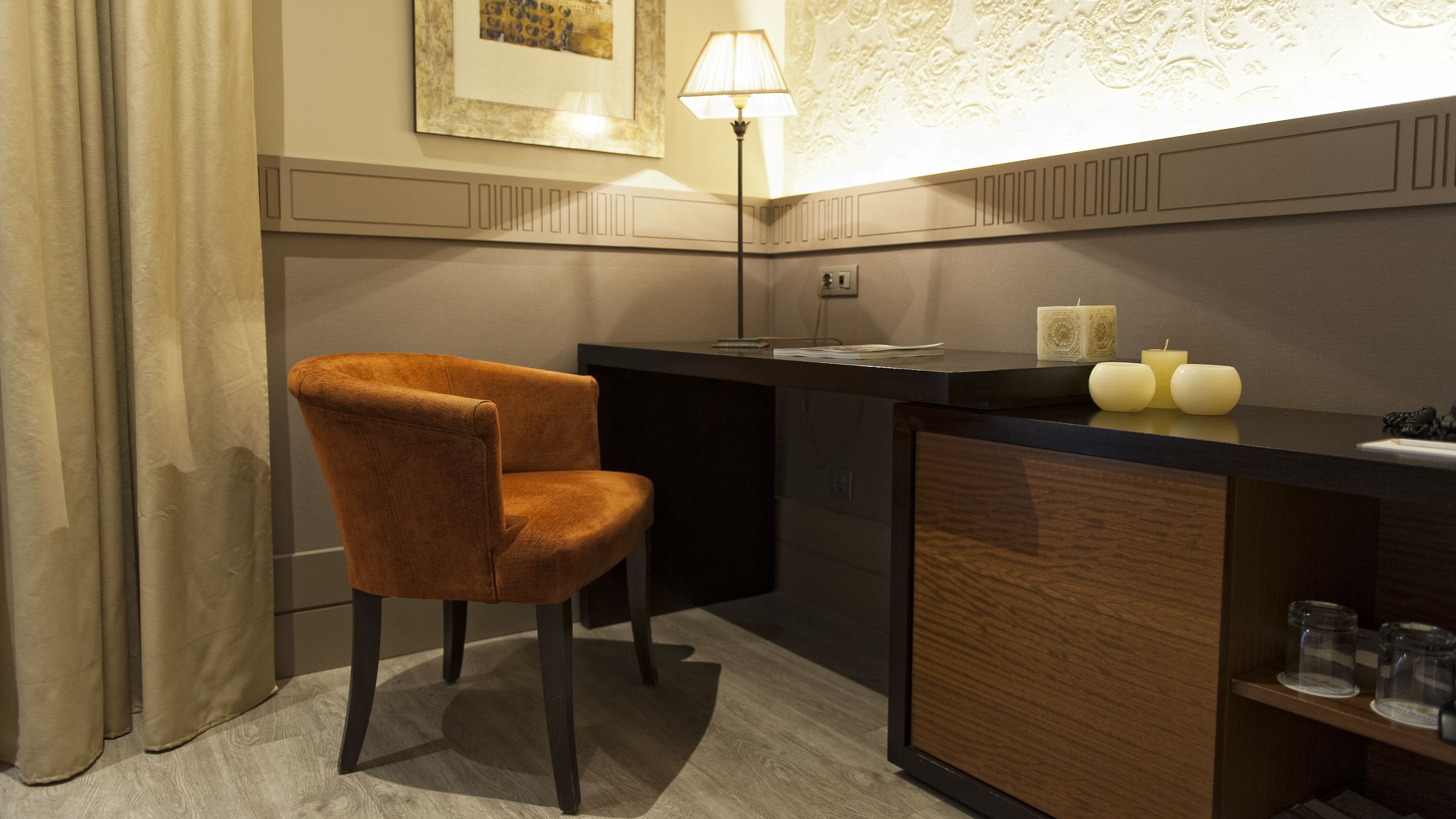 A modern hotel room with a wooden desk, a brown upholstered chair, a lamp, and decorative candles.