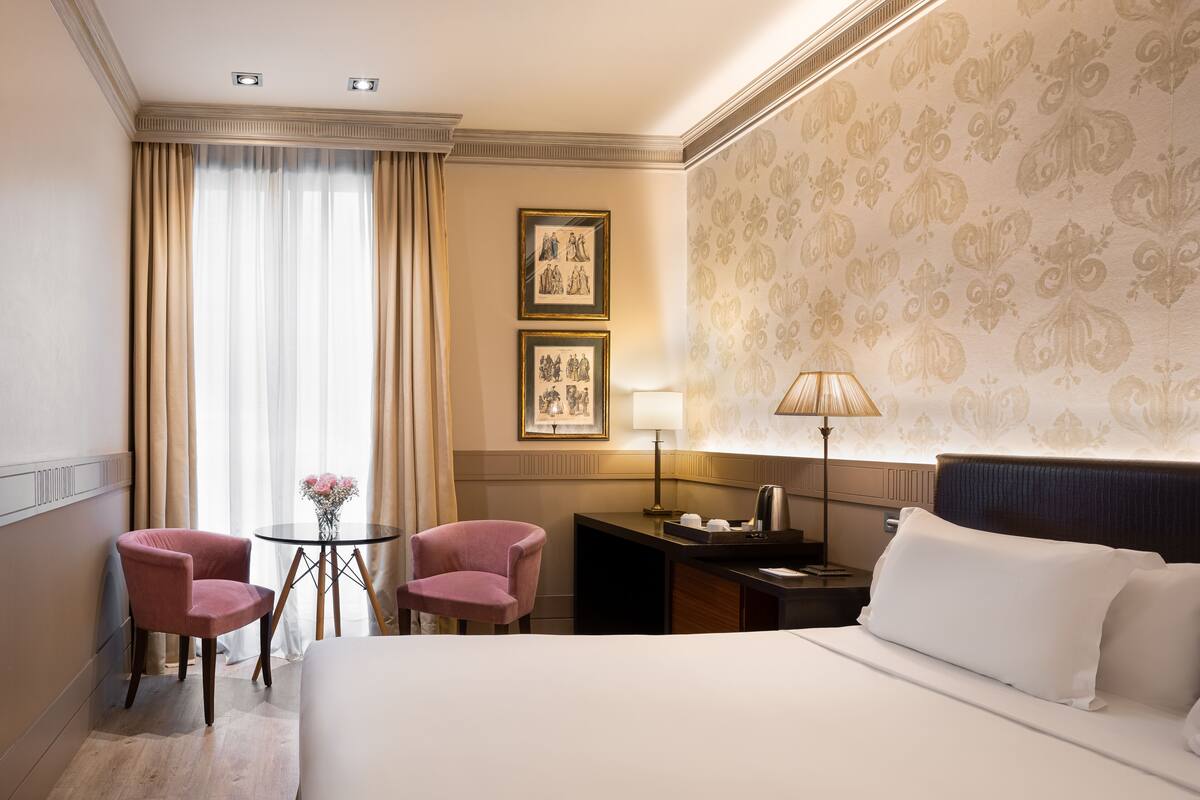 Ein Hotelzimmer mit einem großen Bett, zwei rosafarbenen Stühlen, einem kleinen Tisch mit Blumenvase, einer Lampe und gerahmten Bildern an der Wand.