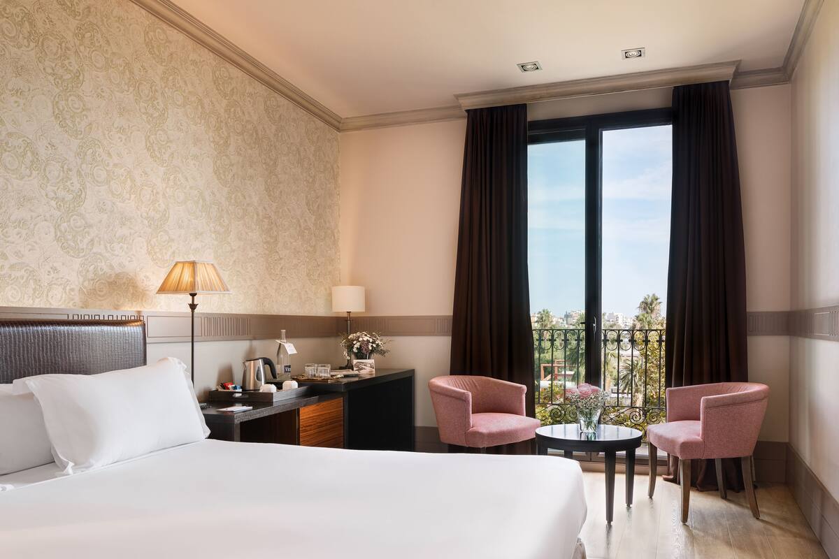 Ein Hotelzimmer mit einem Bett, einem Schreibtisch mit Lampe, zwei rosa Stühlen und Blick auf die Stadt durch große Fenster.