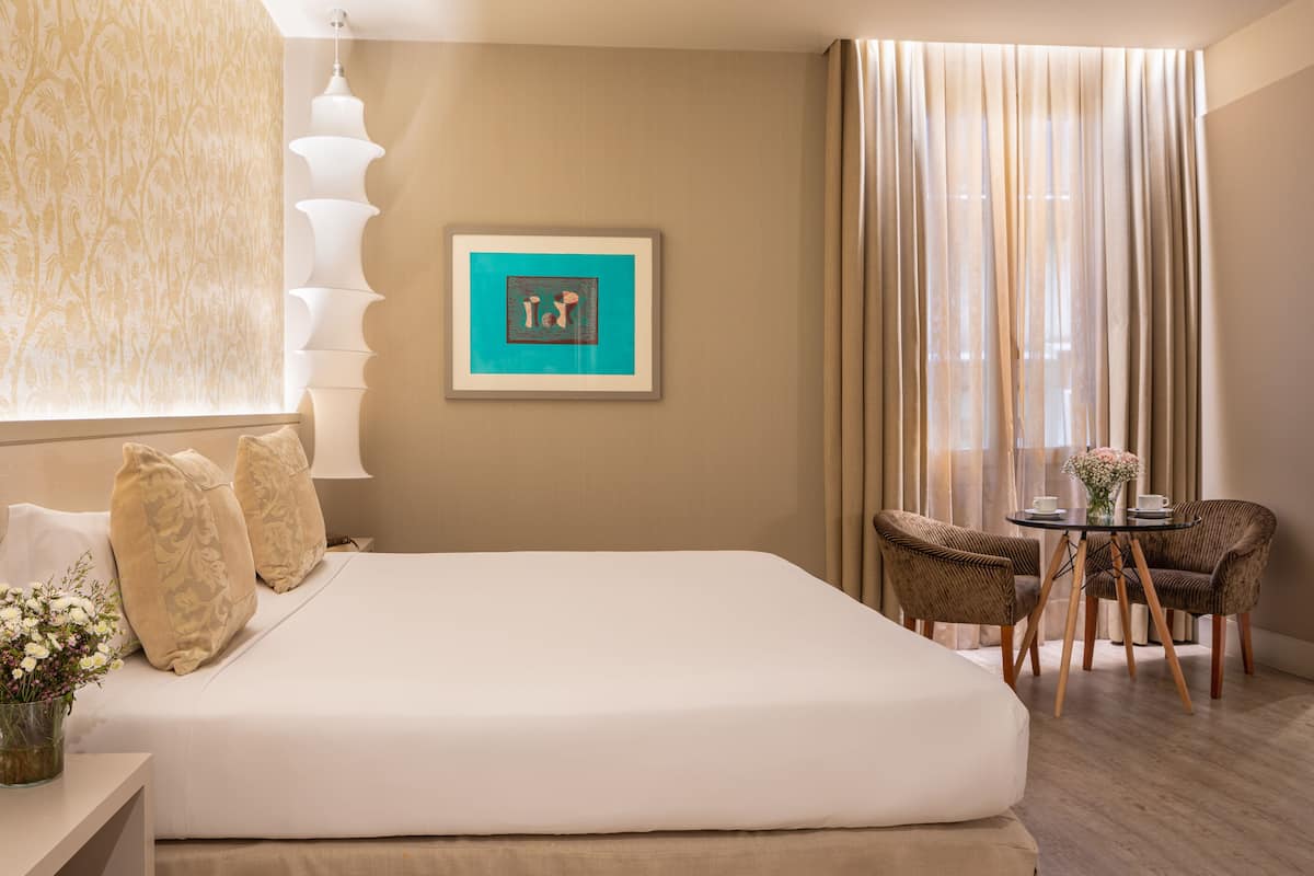 Ein Hotelzimmer mit einem großen Bett, einem kleinen Tisch mit Stühlen, einer Vase mit Blumen und einem gerahmten Bild an der Wand.