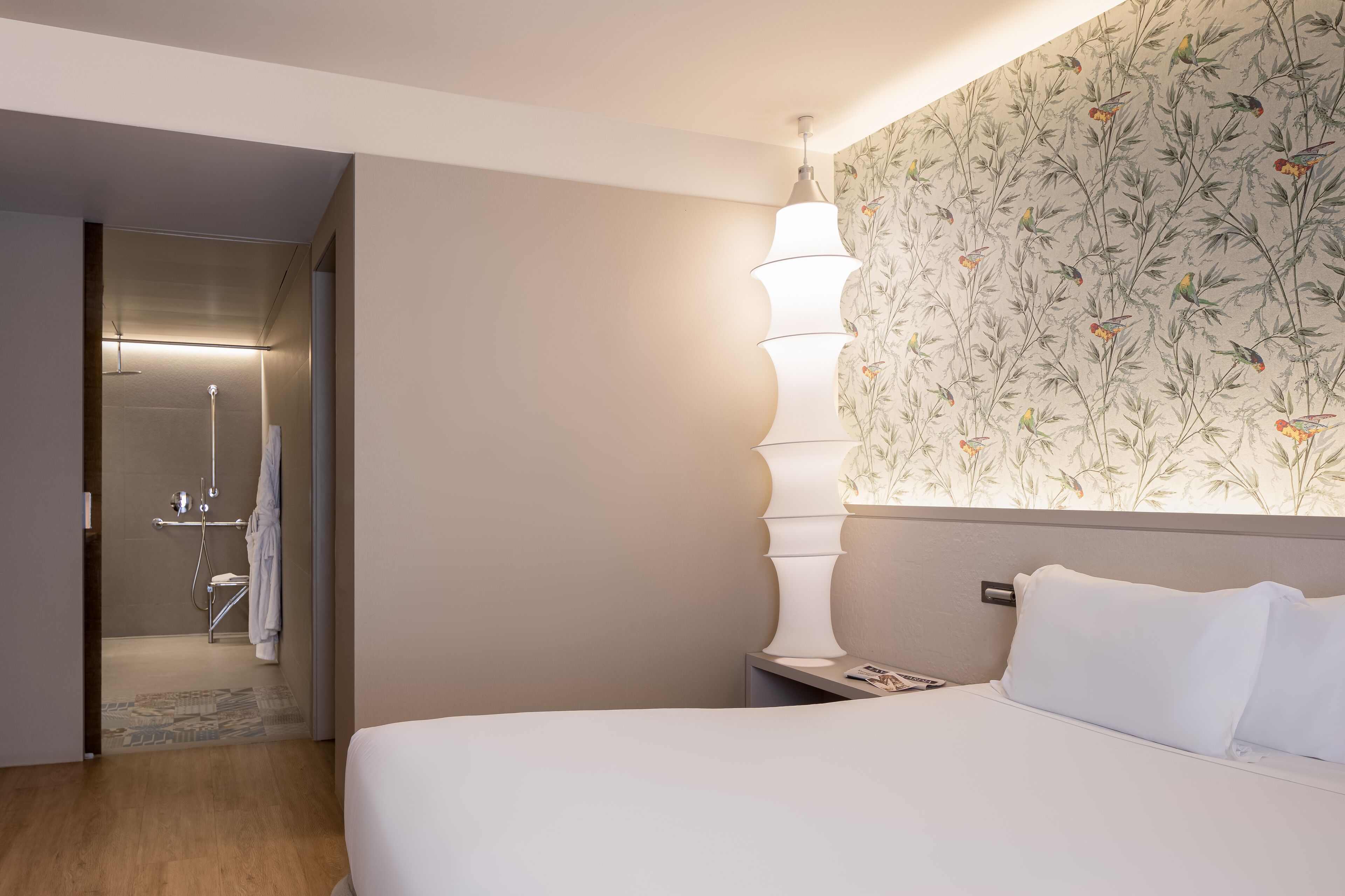 Hotelkamer met een bed, nachtkastje, wandlamp en een zichtbare badkamer door een open deur.