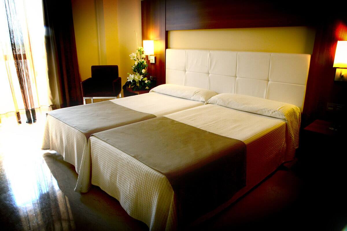 A hotel room with two beds, a nightstand with a lamp, a chair, and a window with curtains.