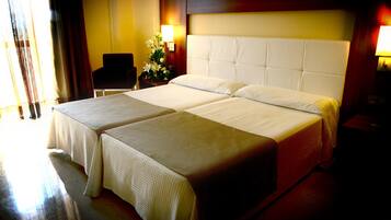 Habitación de hotel con dos camas, una mesita de noche con lámpara, una silla y un ventanal con cortinas.
