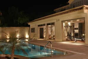 An outdoor swimming pool area with a palm tree, a covered patio, and a building with glass doors and windows. - Hotel Spa Jardines de Lorca (Lorca)