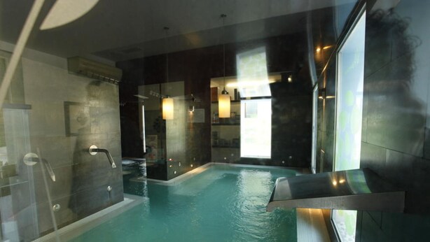 A modern indoor swimming pool with clear glass walls, a built-in bench, and a view of the outside.