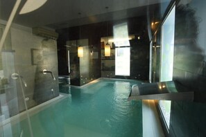 A modern indoor swimming pool with clear glass walls, a built-in bench, and a view of the outside. - Hotel Spa Jardines de Lorca (Lorca)