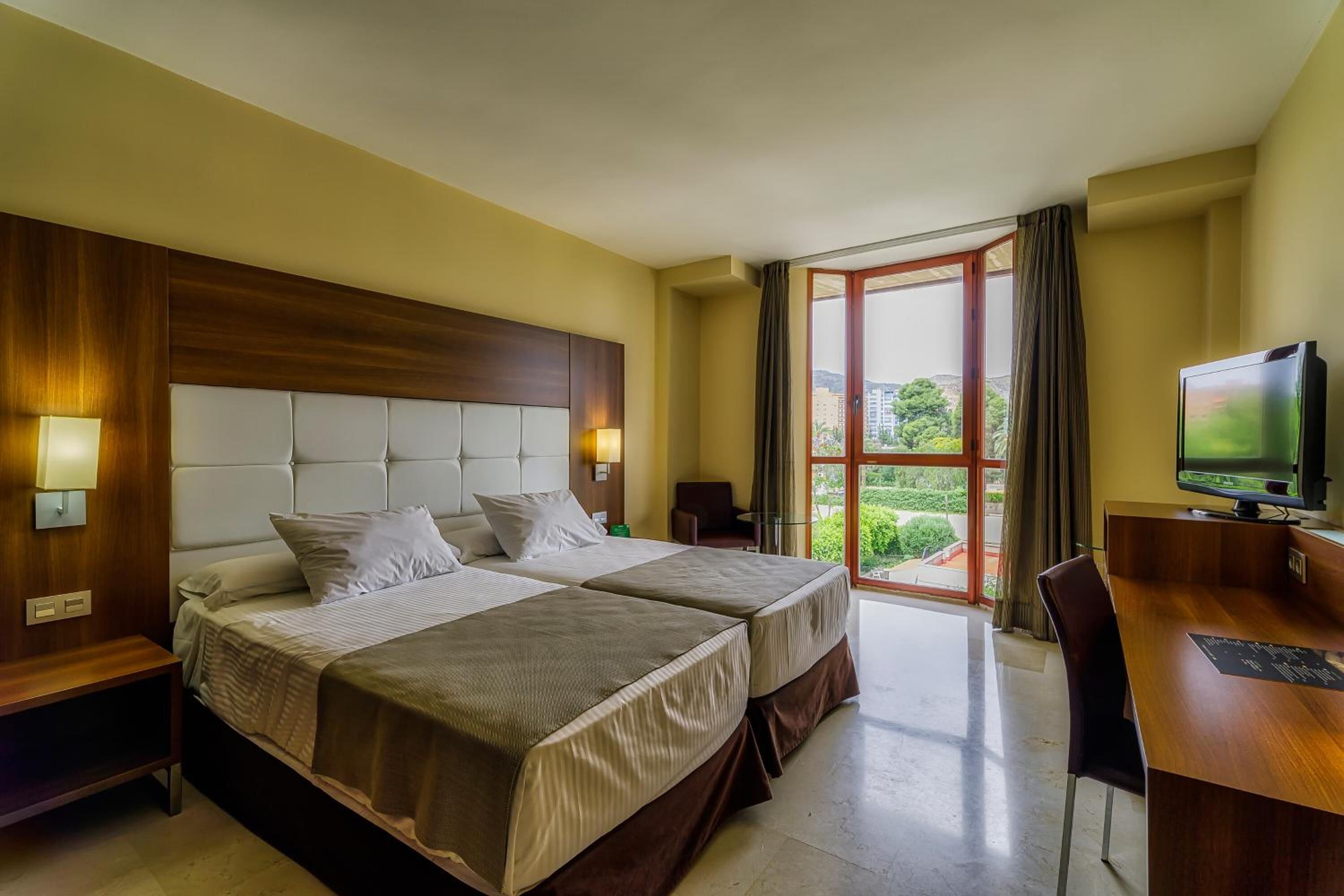 Ein Hotelzimmer mit zwei Betten, einem Schreibtisch, einem Fernseher und einem großen Fenster mit Blick auf Grünflächen.