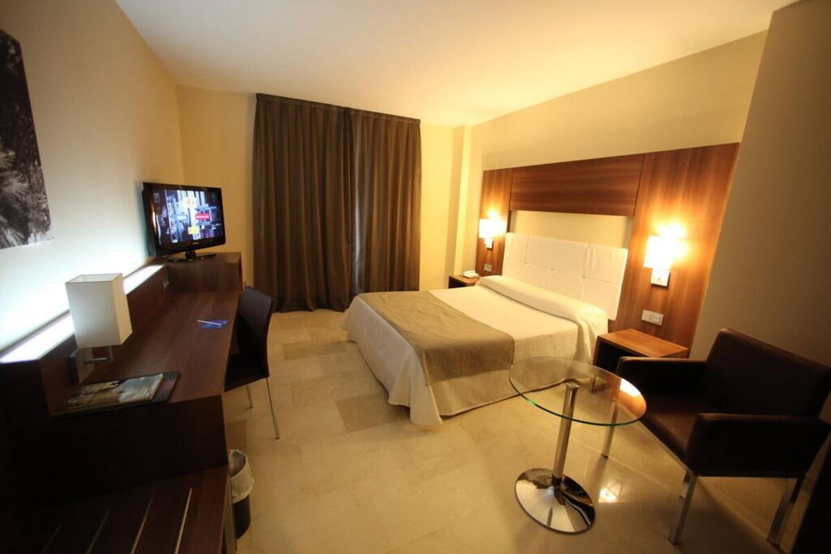 A hotel room with a bed, a desk, a television, a glass table, and two bedside lamps.