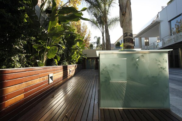 A wooden deck with a glass enclosure, surrounded by lush greenery and a modern building.