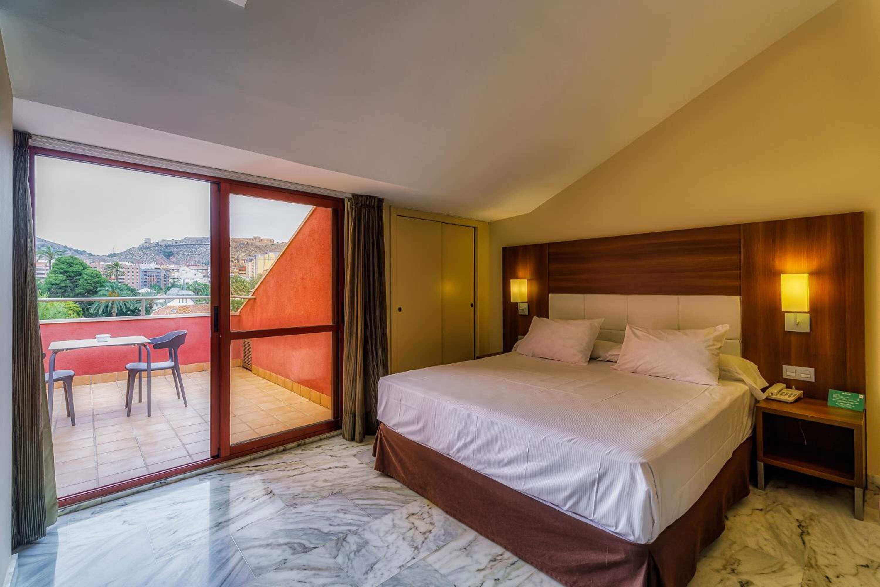 A hotel room with a bed, a nightstand, a desk with chairs, a sliding glass door leading to an outdoor area, and a city view.