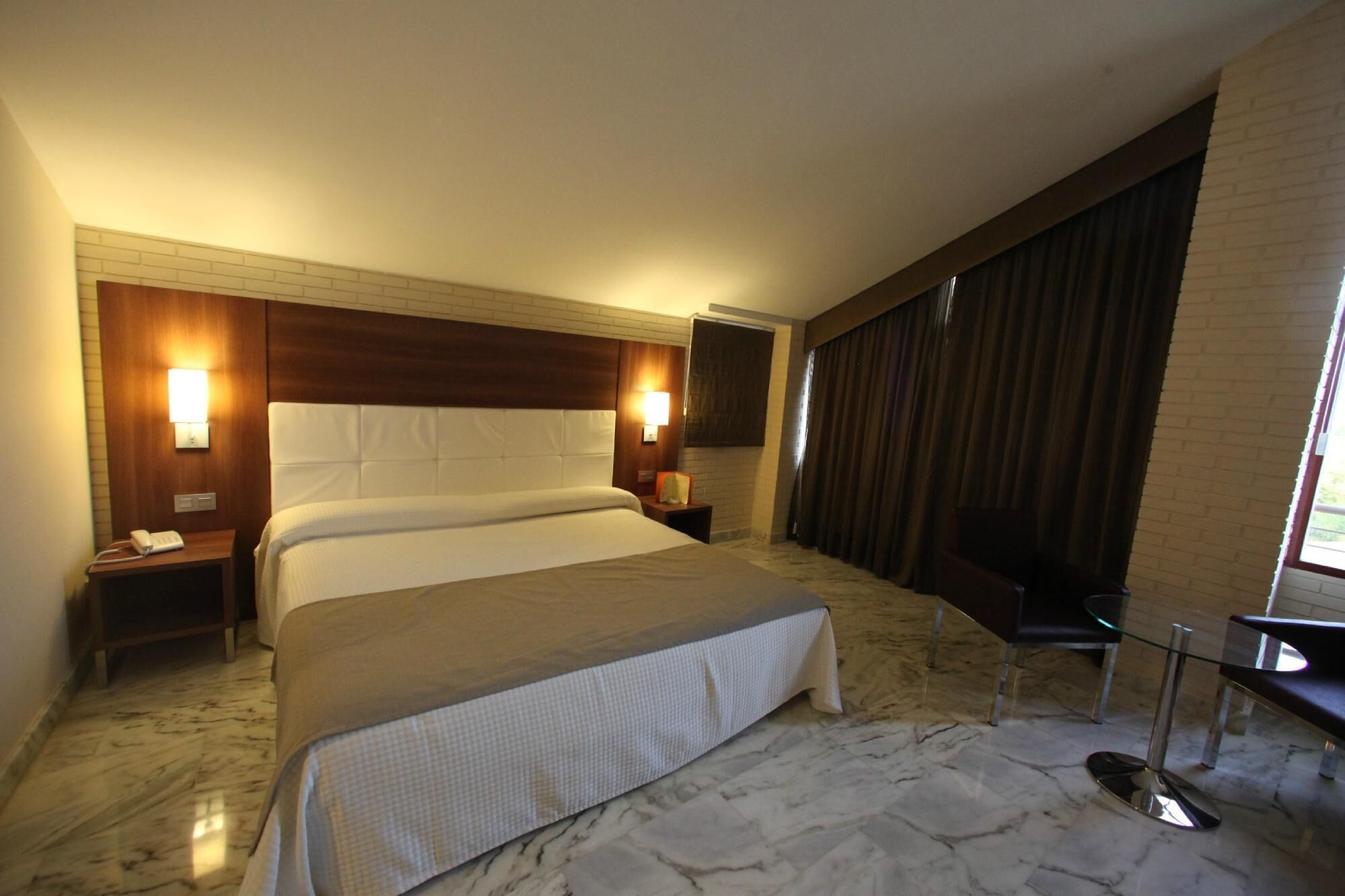 A hotel room with a large bed, two bedside tables, a nightstand, a glass table, and a view of the outdoors.