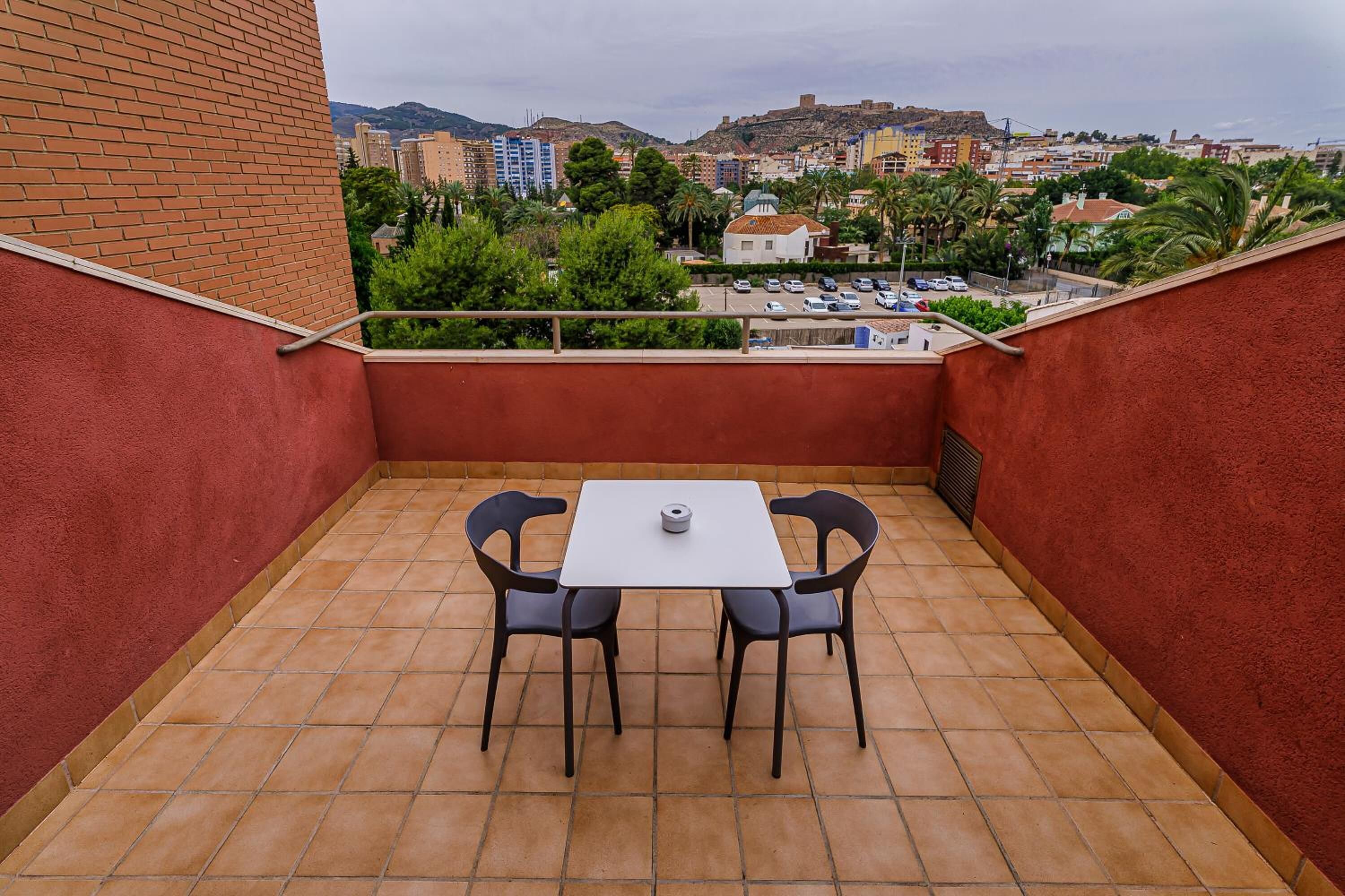 A small balcony with a table and two chairs, overlooking a cityscape with buildings and palm trees.