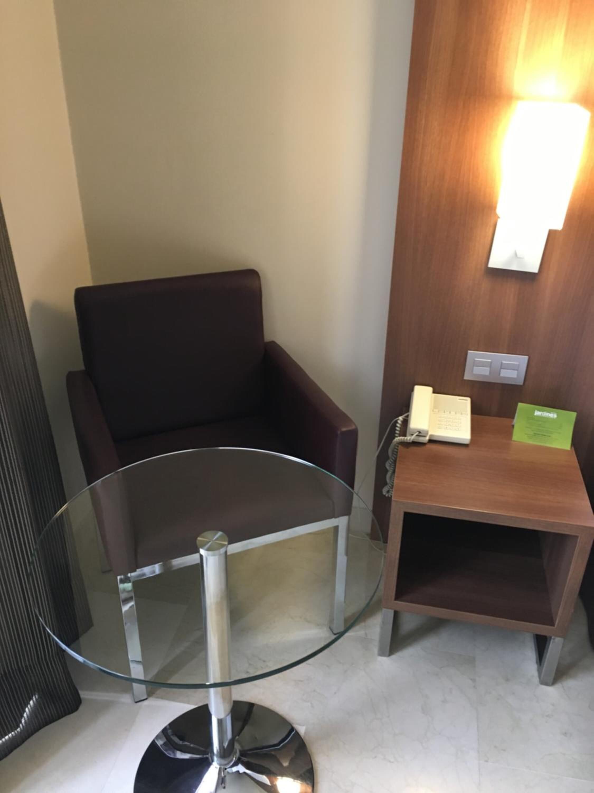 A modern room with a glass-top table, a brown leather chair, a wooden side table with a phone and a green card, and a wall-mounted light fixture.
