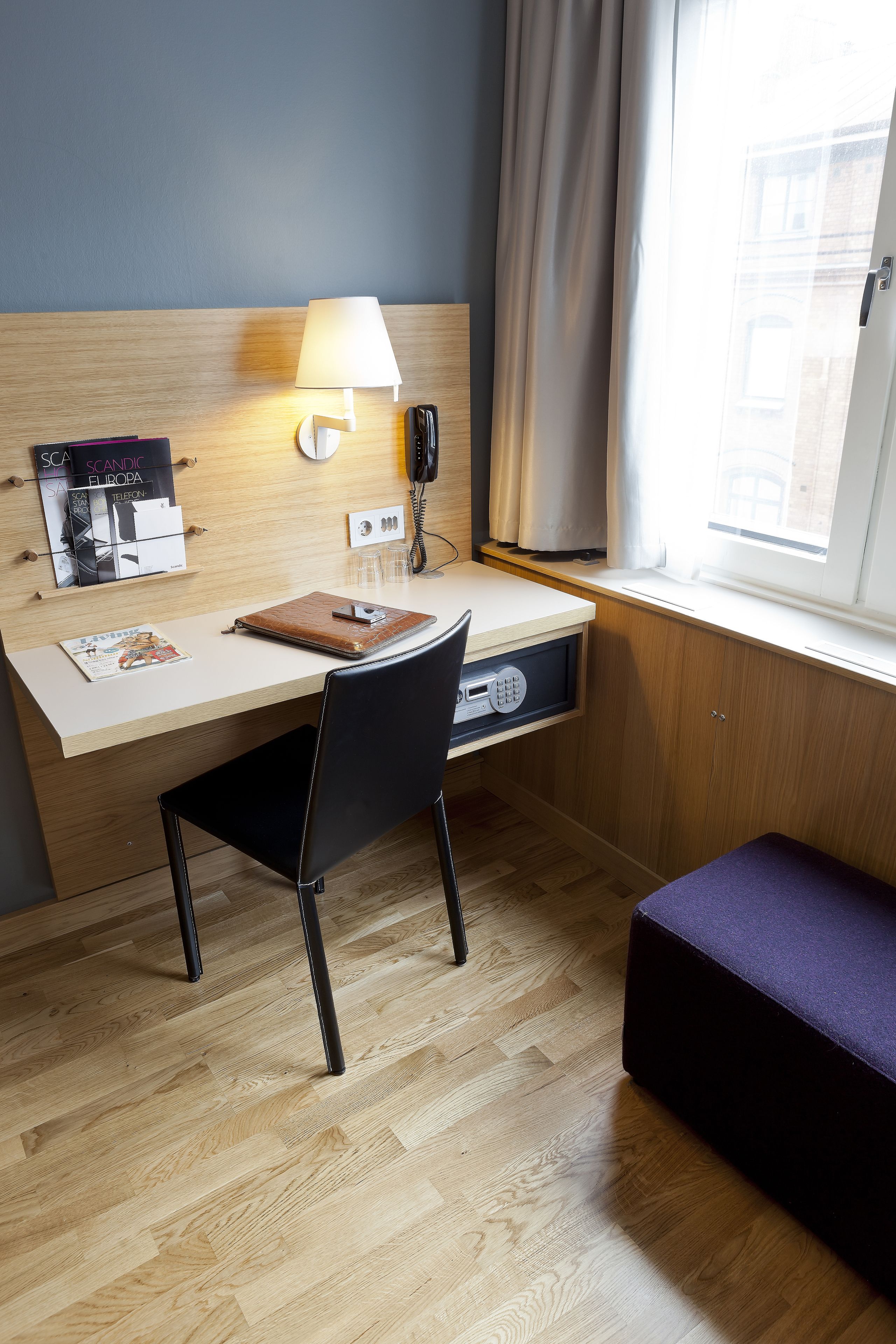 Una habitación de hotel con un escritorio de madera, una silla, una lámpara, un teléfono y una ventana con cortinas.