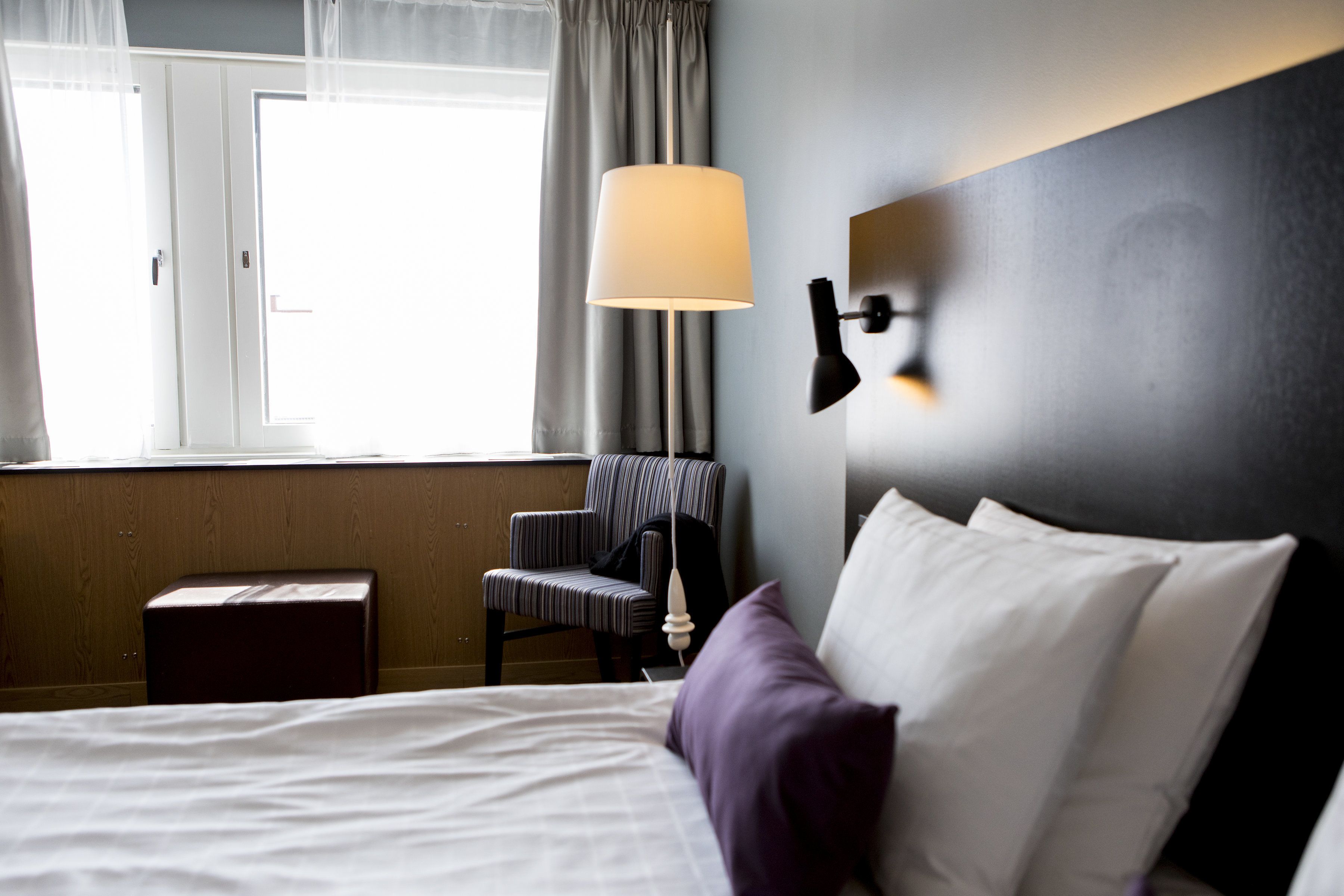 A hotel room with a bed, a chair, a lamp, and a window with curtains.