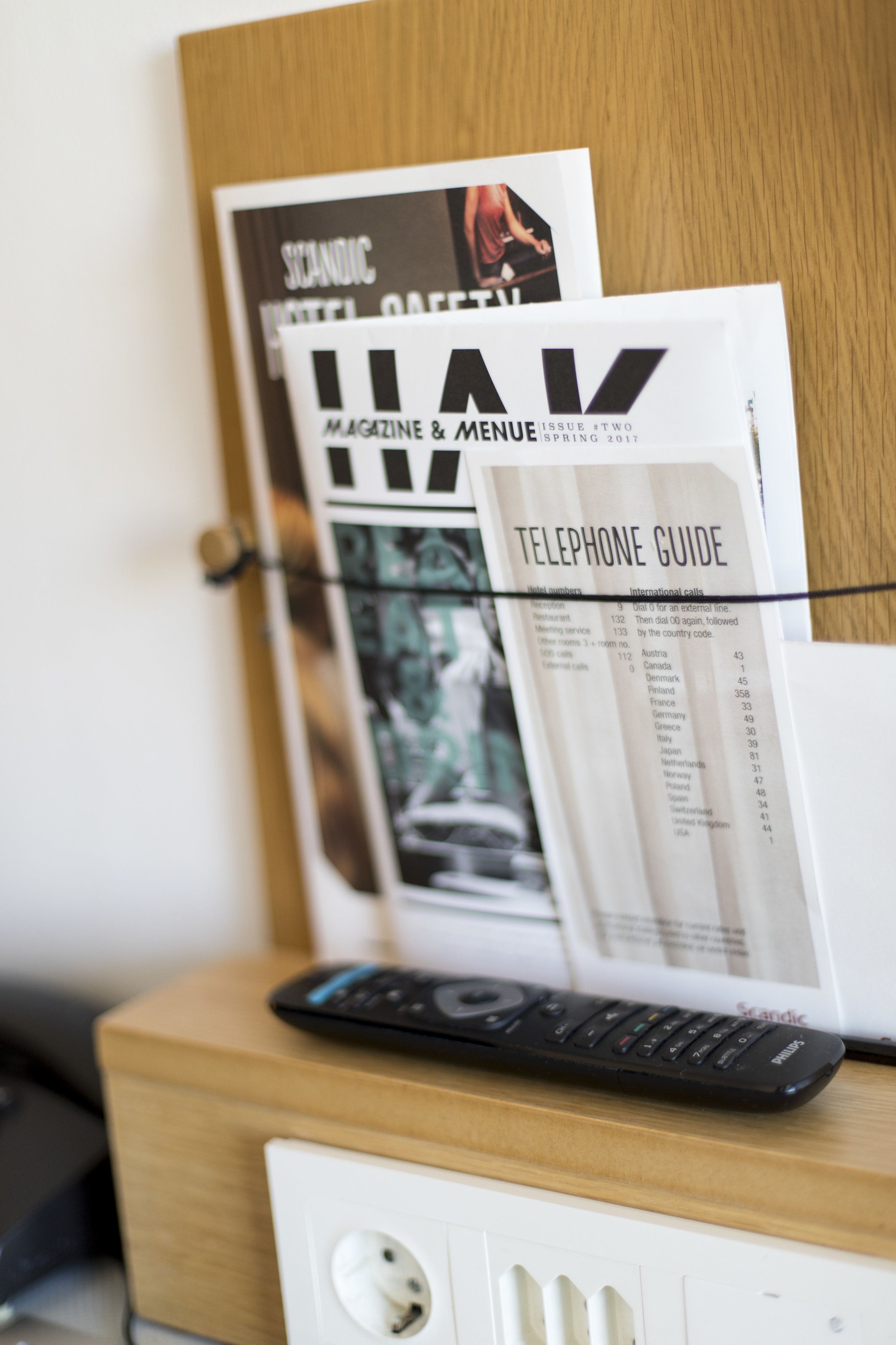A wooden desk with a remote control, a telephone guide, and a magazine on the wall.