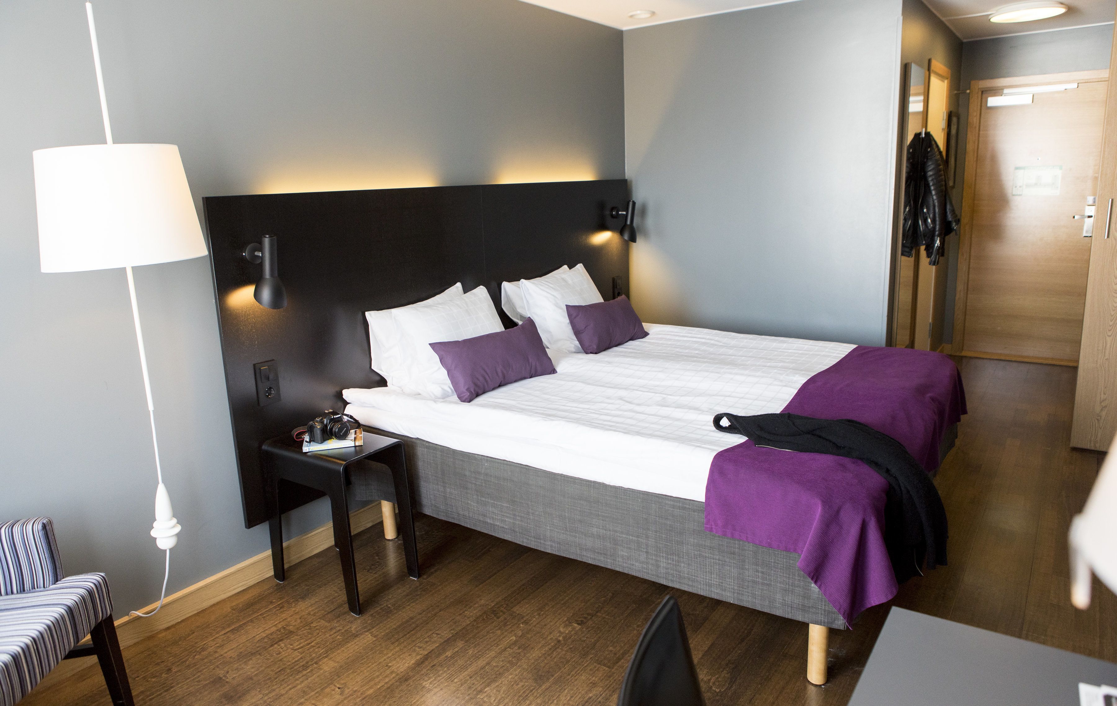 Una cama bien hecha con ropa de cama blanca y morada, una mesita de noche con una lámpara y un perchero con una chaqueta.