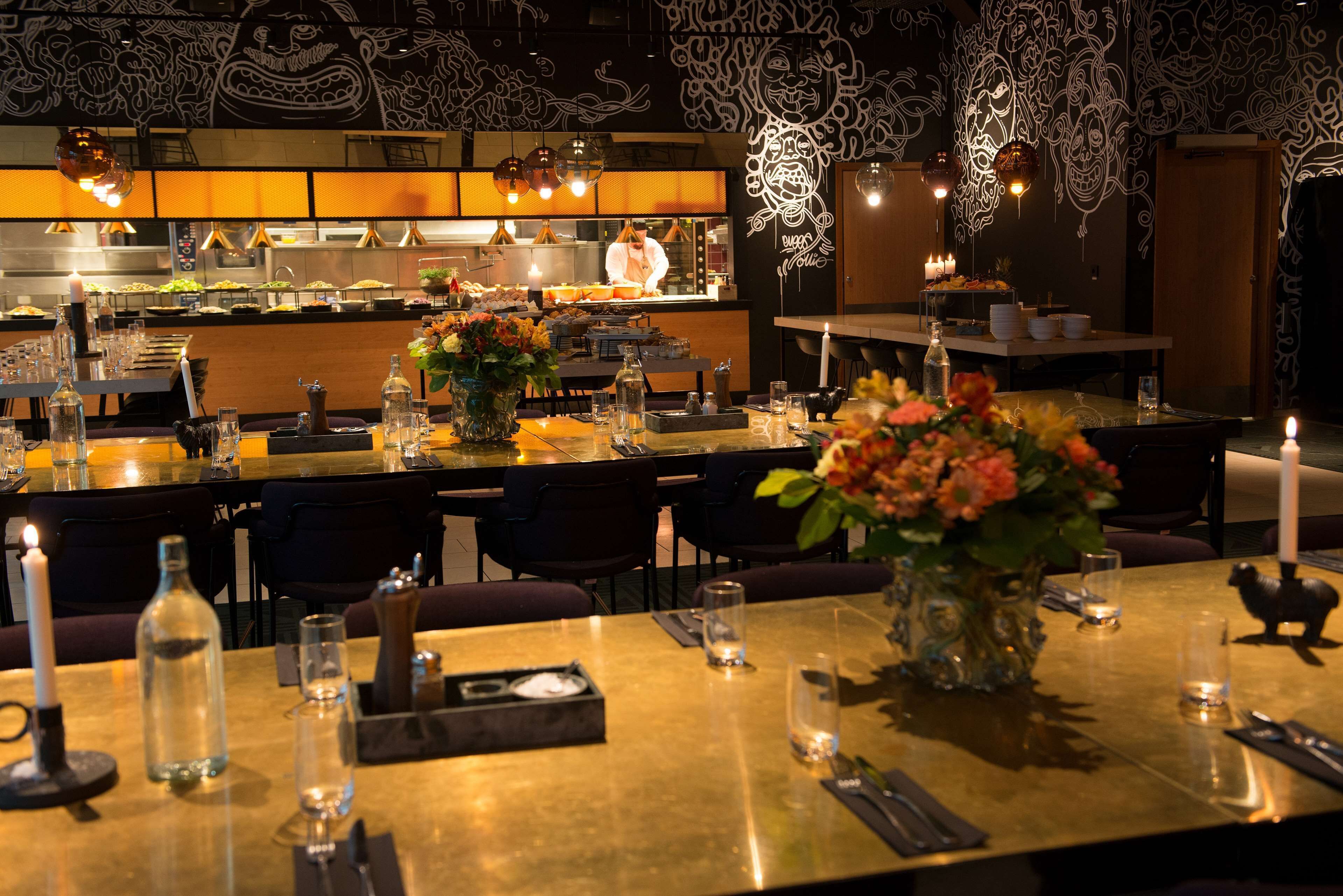 Un restaurante con mesas dispuestas para comer, un chef al fondo y decoración mural.