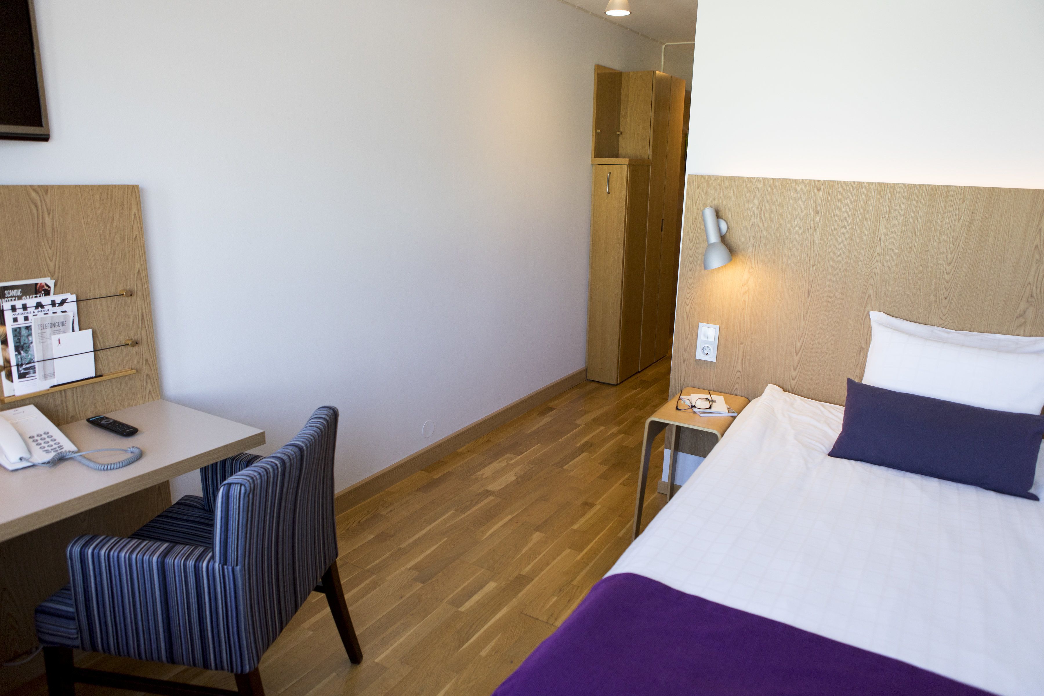 Ein Hotelzimmer mit Bett, Schreibtisch mit Telefon, Stuhl, Nachttisch mit Lampe und Holzschrank.