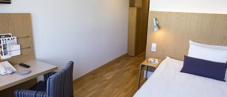 Ein Hotelzimmer mit Bett, Schreibtisch mit Telefon, Stuhl, Nachttisch mit Lampe und Holzschrank.