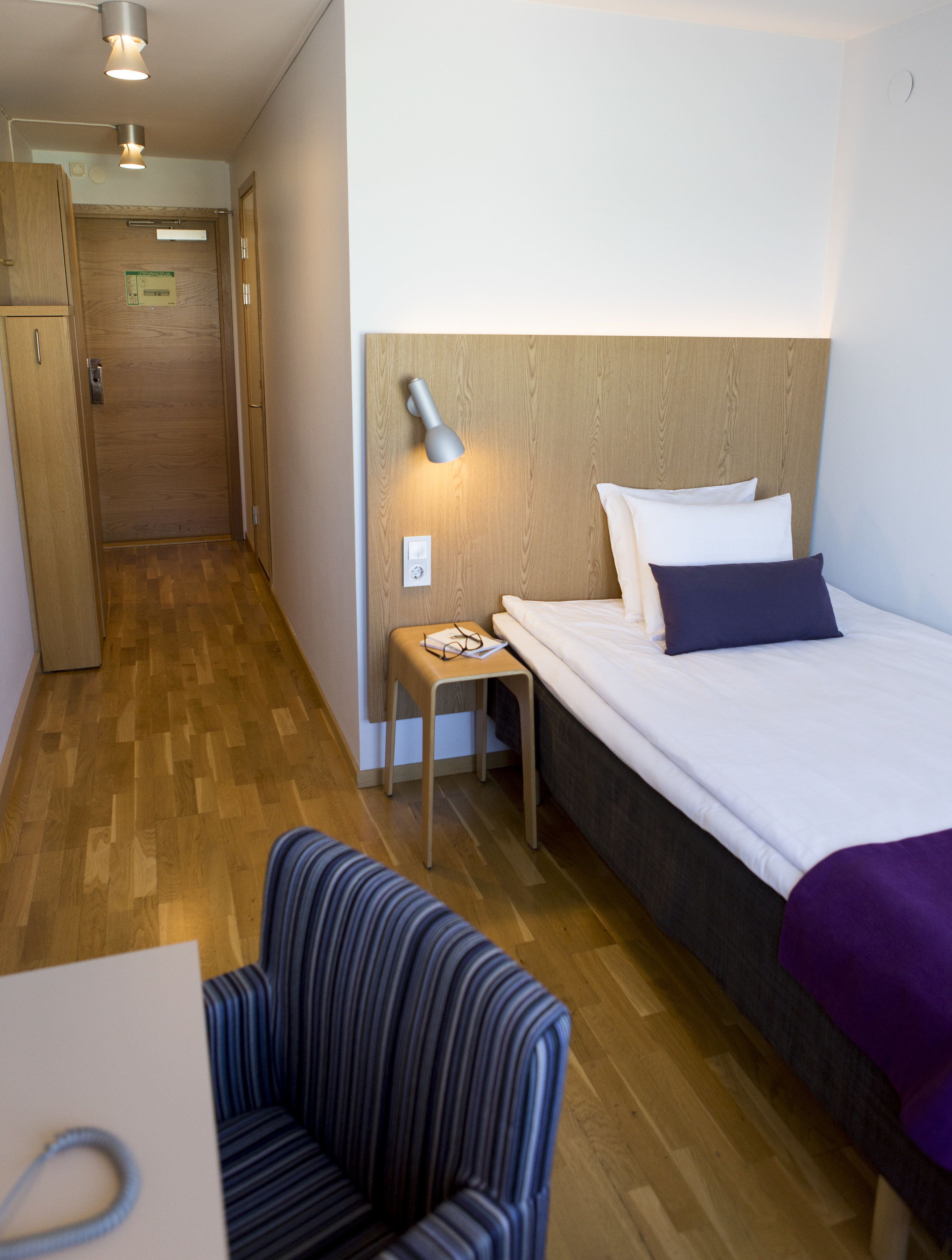 A hotel room with a wooden floor, a bed with white and dark pillows, a bedside table with glasses, a chair, and a wooden headboard.