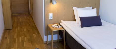 A hotel room with a wooden floor, a bed with white and dark pillows, a bedside table with glasses, a chair, and a wooden headboard.