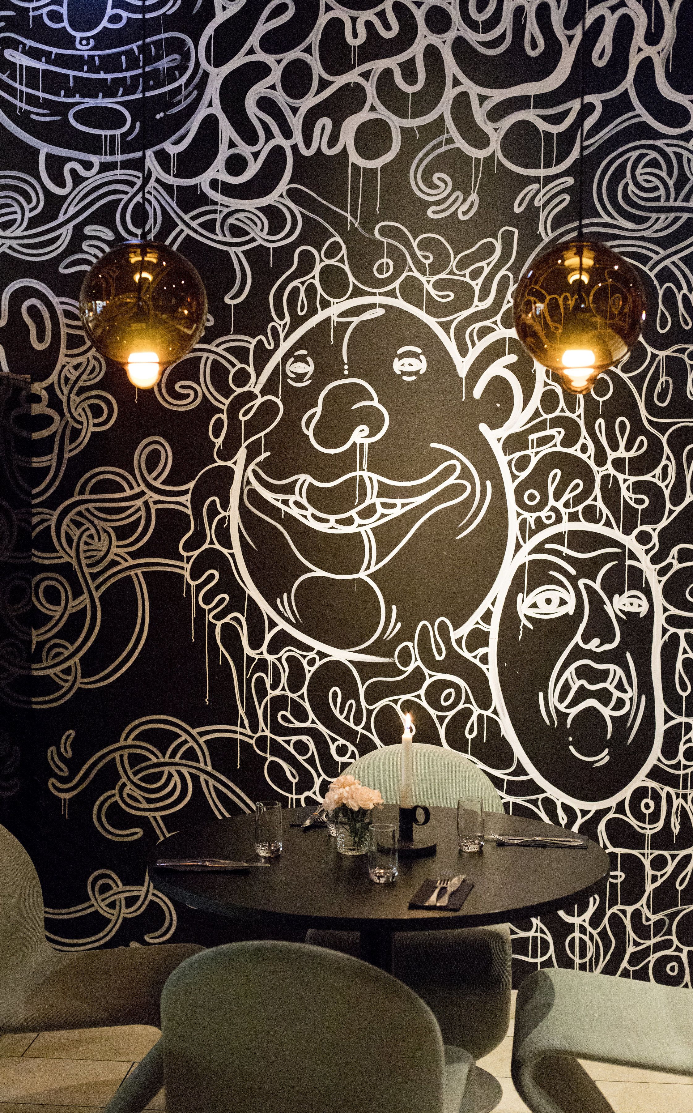 Un coin repas avec une table dressée pour deux personnes, un mur décoratif orné de motifs abstraits et deux lampes suspendues.