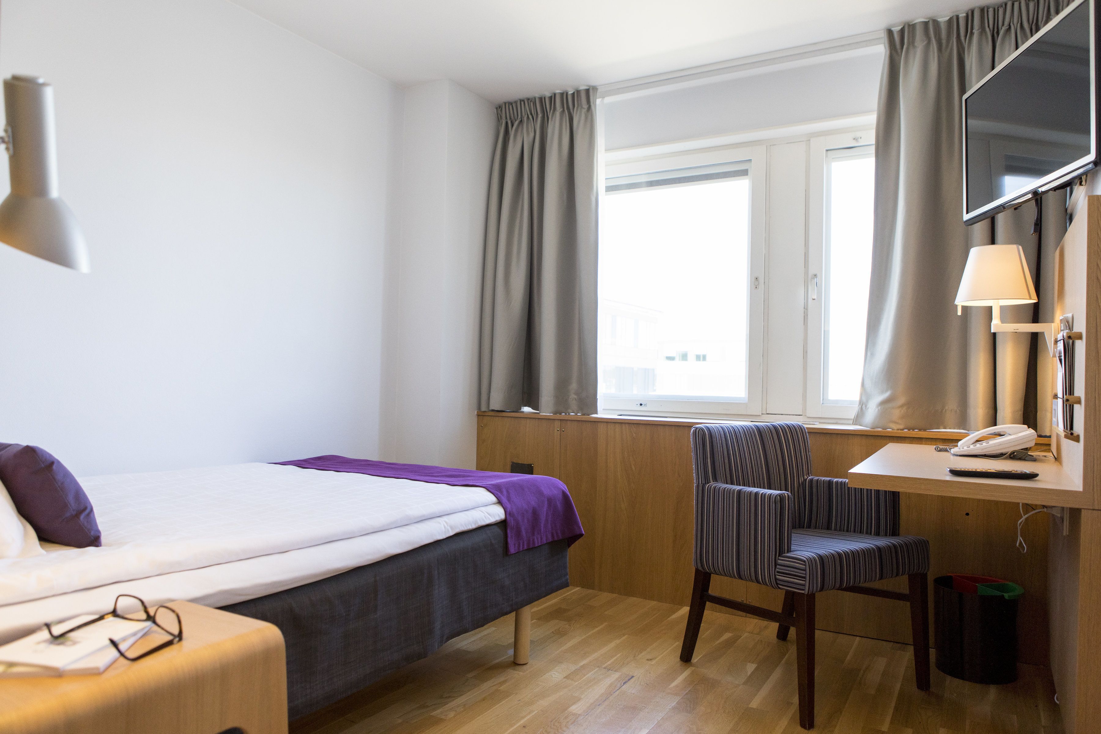 Habitación de hotel con cama, escritorio, silla, televisor y ventana con cortinas.