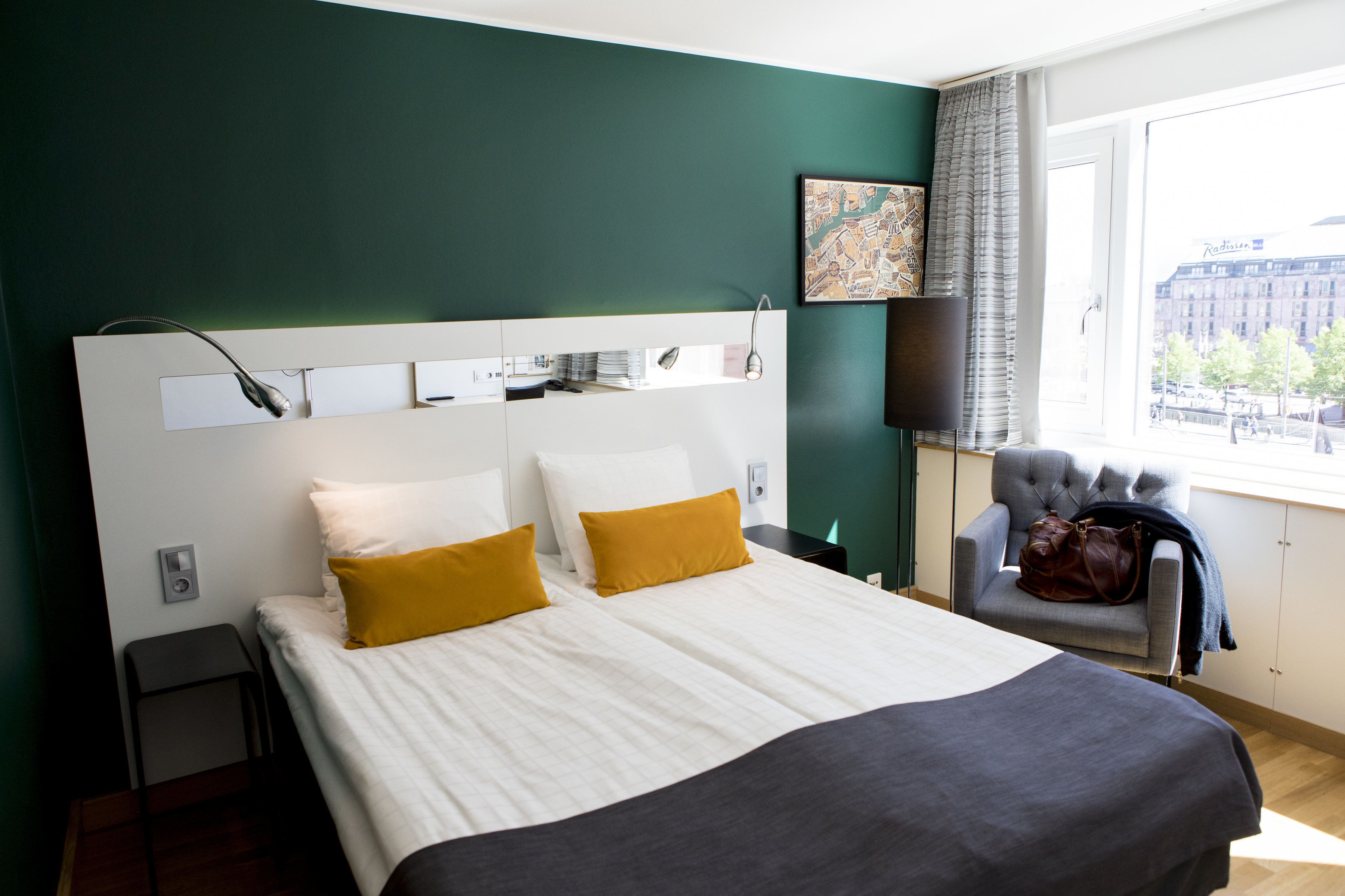 A hotel room with a large bed, a chair, a desk, and a window with a city view.