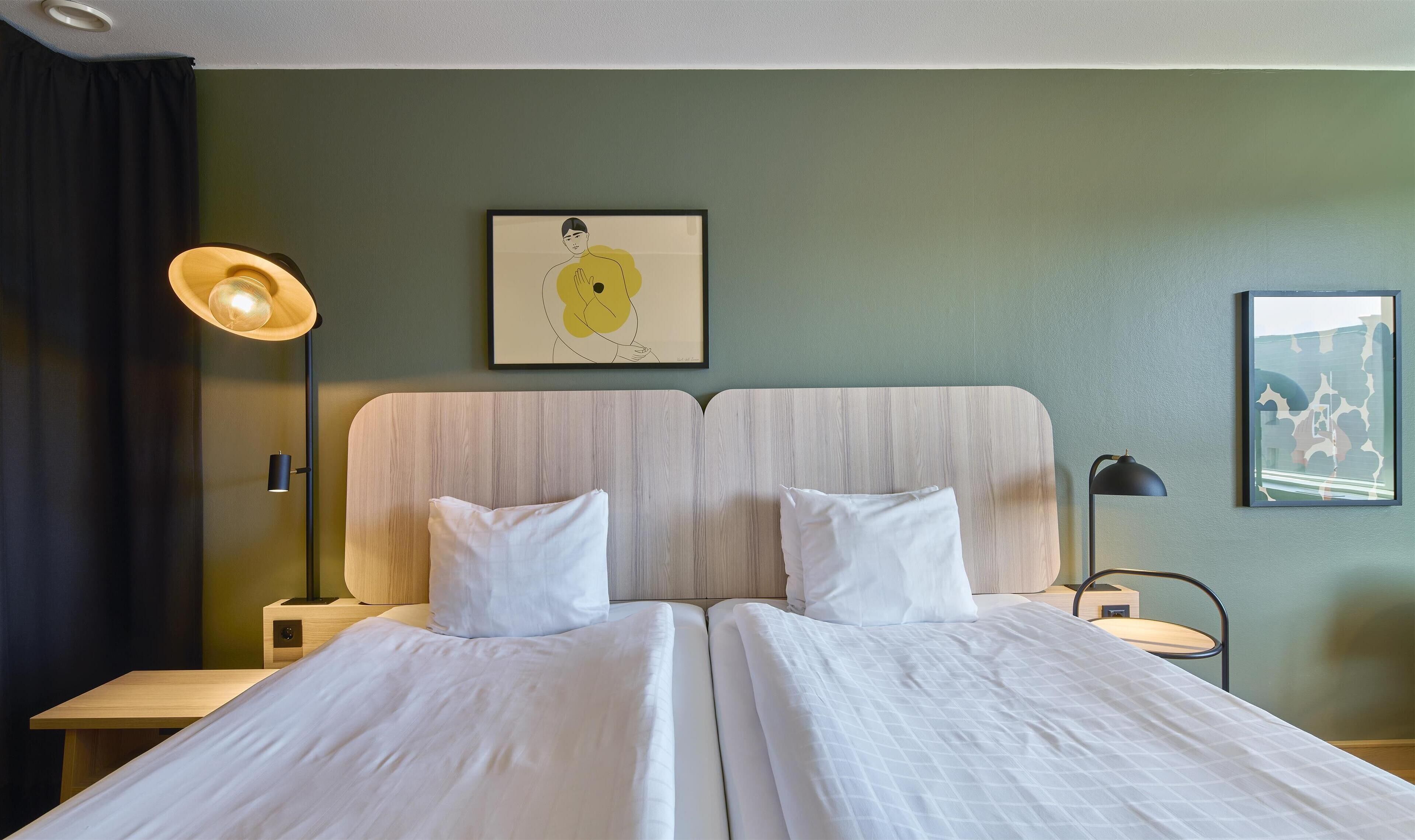Cama doble con sábanas blancas, cabecera de madera, mesita de noche con lámpara y un cuadro enmarcado en la pared.
