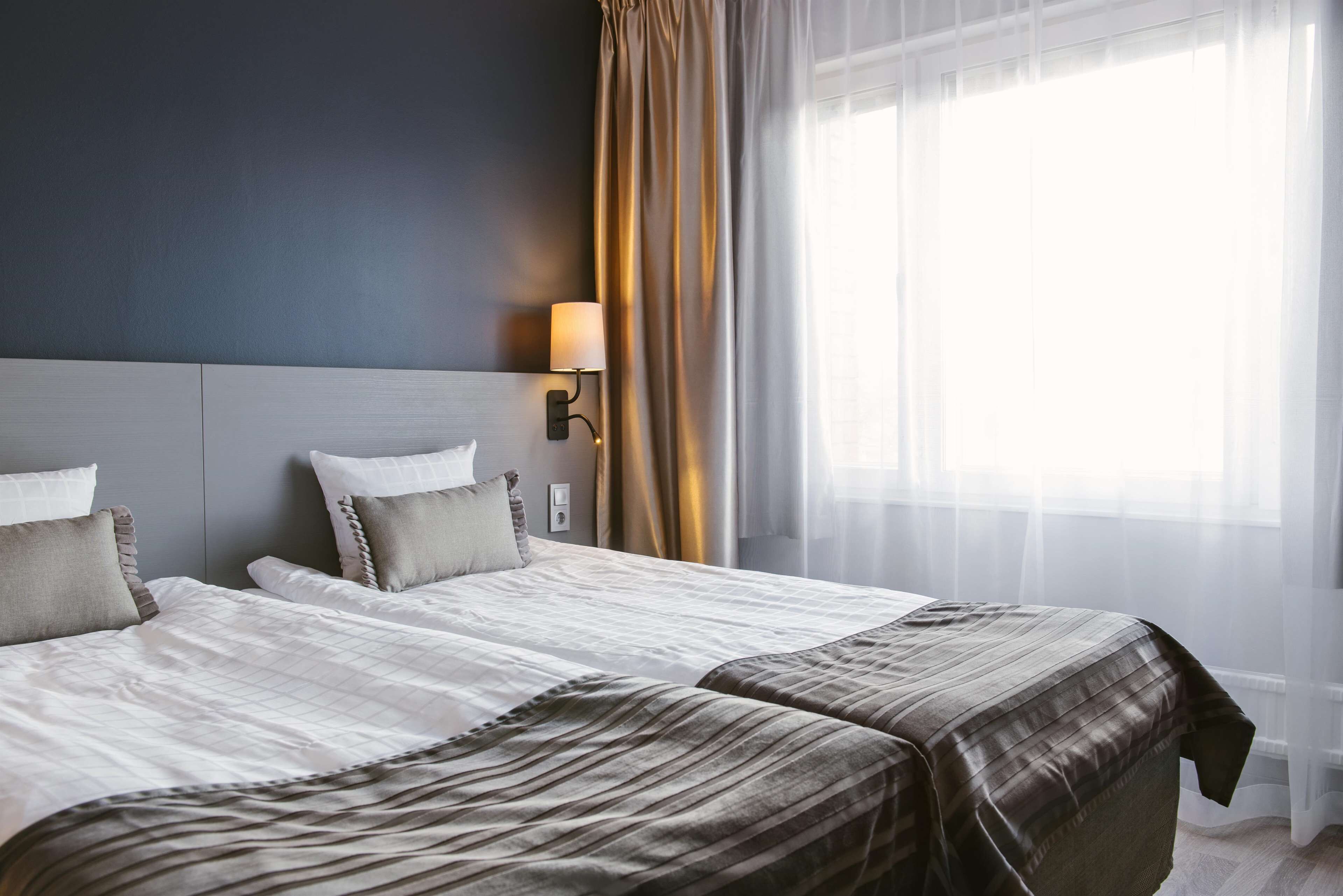 Habitación de hotel con dos camas, cabecero, lámpara de pared y ventana con cortinas traslúcidas.