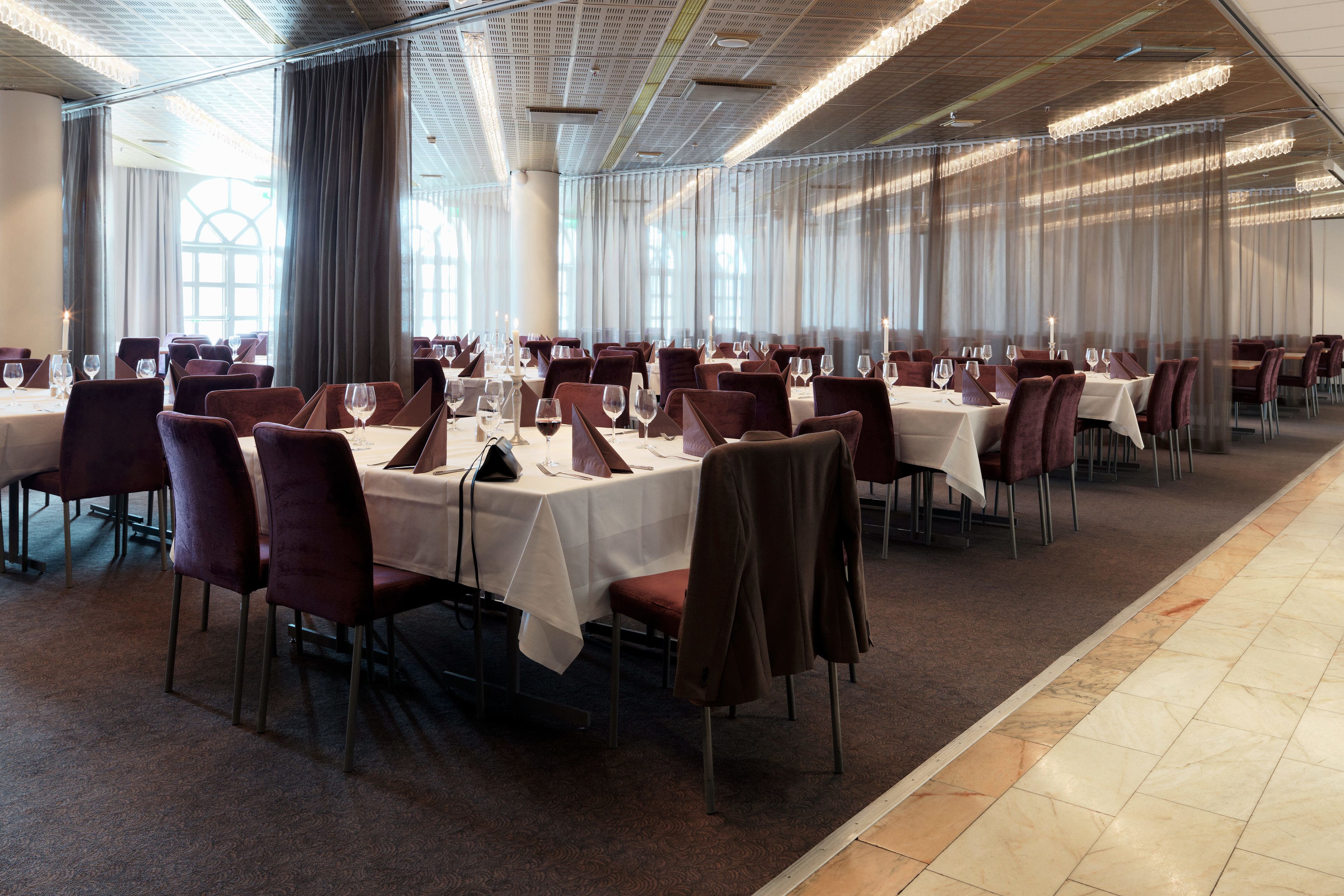 A banquet hall with tables set for dining, large windows with curtains, and a patterned carpet.