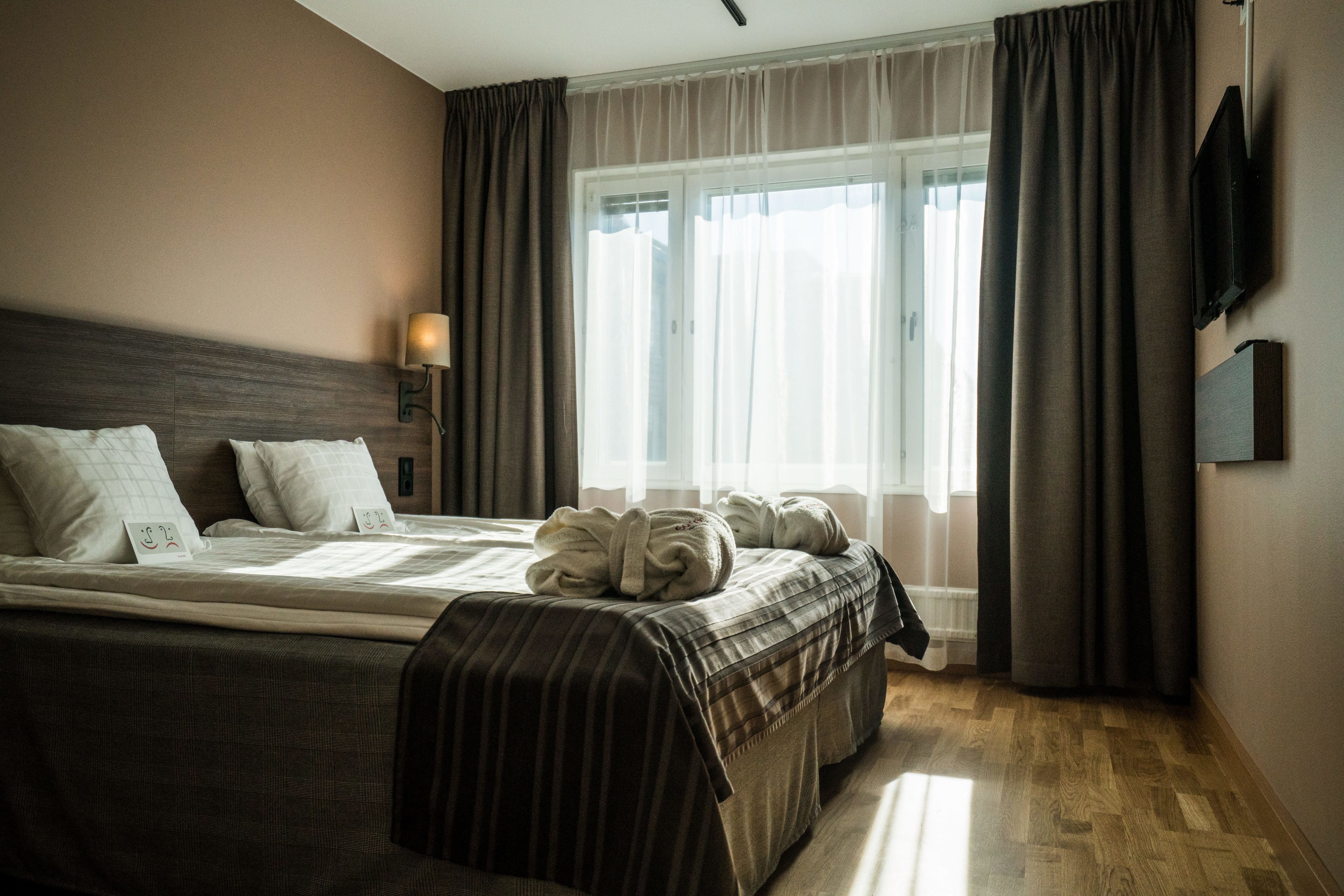 A hotel room with a large bed, a bedside lamp, a TV mounted on the wall, and a window with curtains.