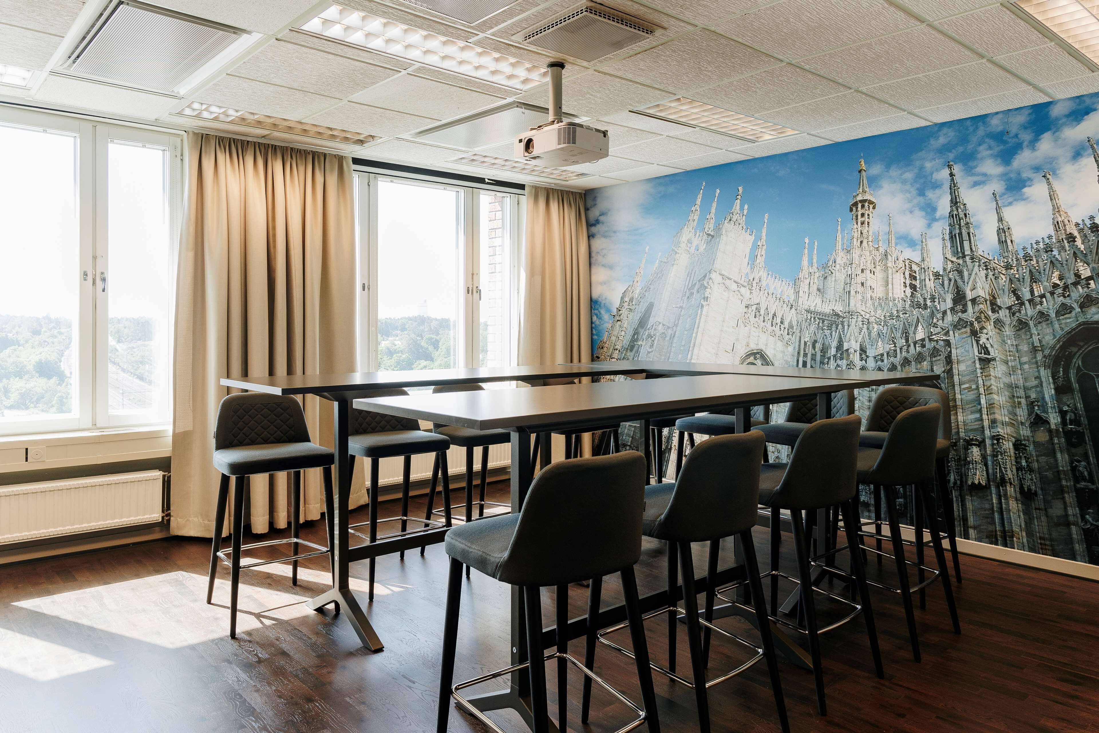 Una sala de conferències amb una taula gran i diverses cadires, un projector al sostre, grans finestres amb cortines i un mural d'una catedral.