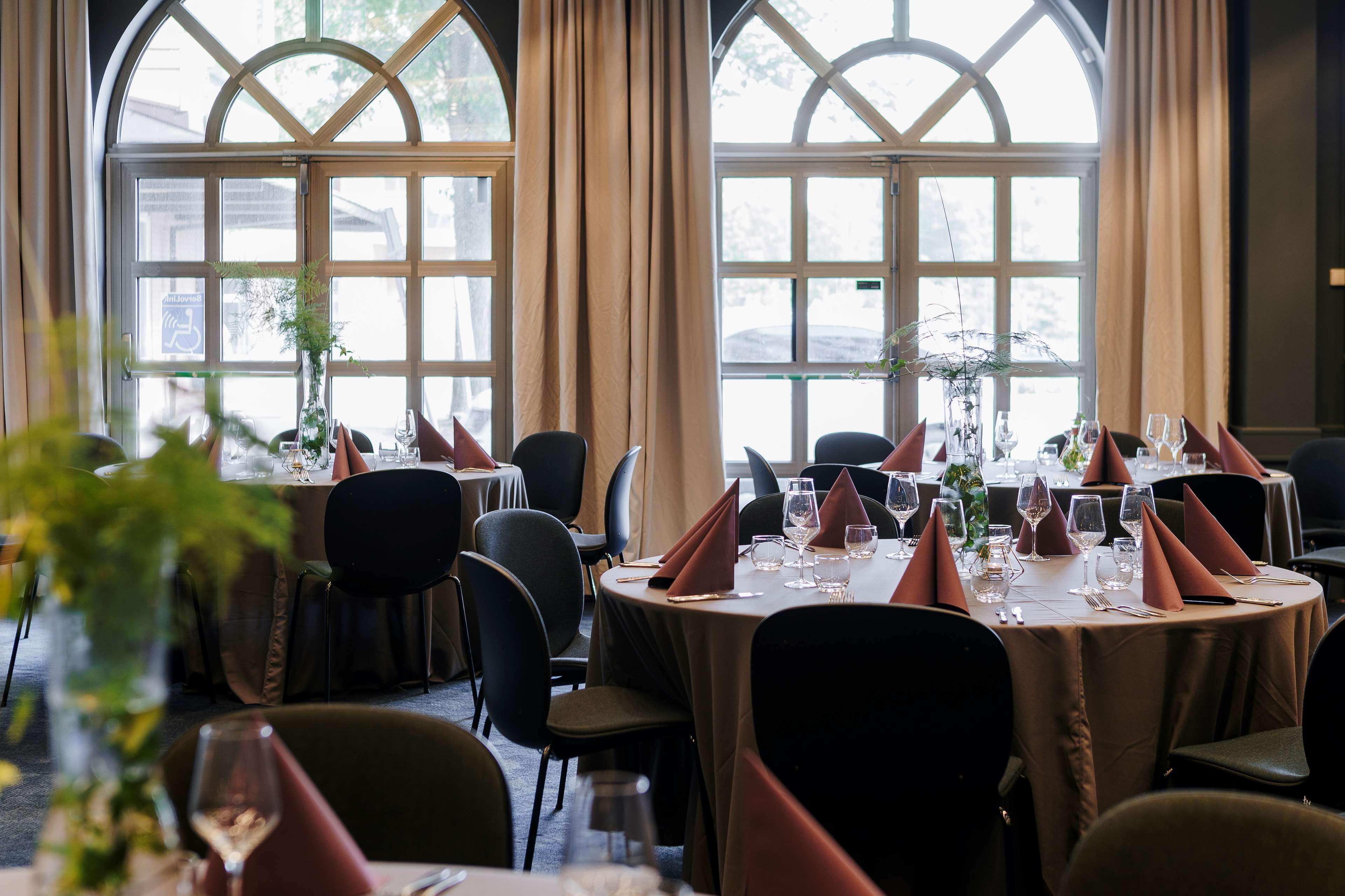 A restaurant with round tables set for dining, large windows with curtains, and indoor plants.