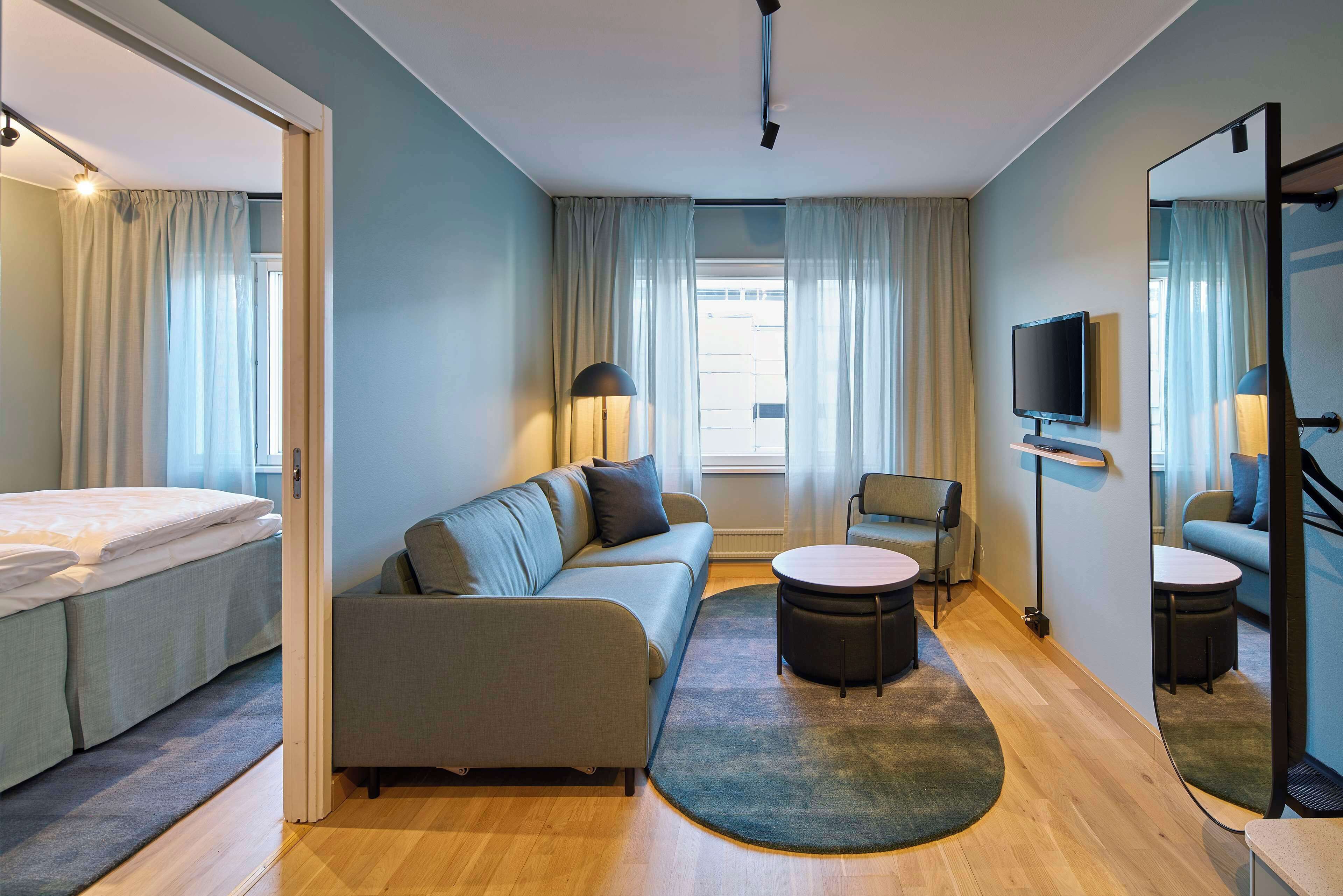 A modern hotel room with a sofa, a round coffee table, a small chair, a bed, and a TV.