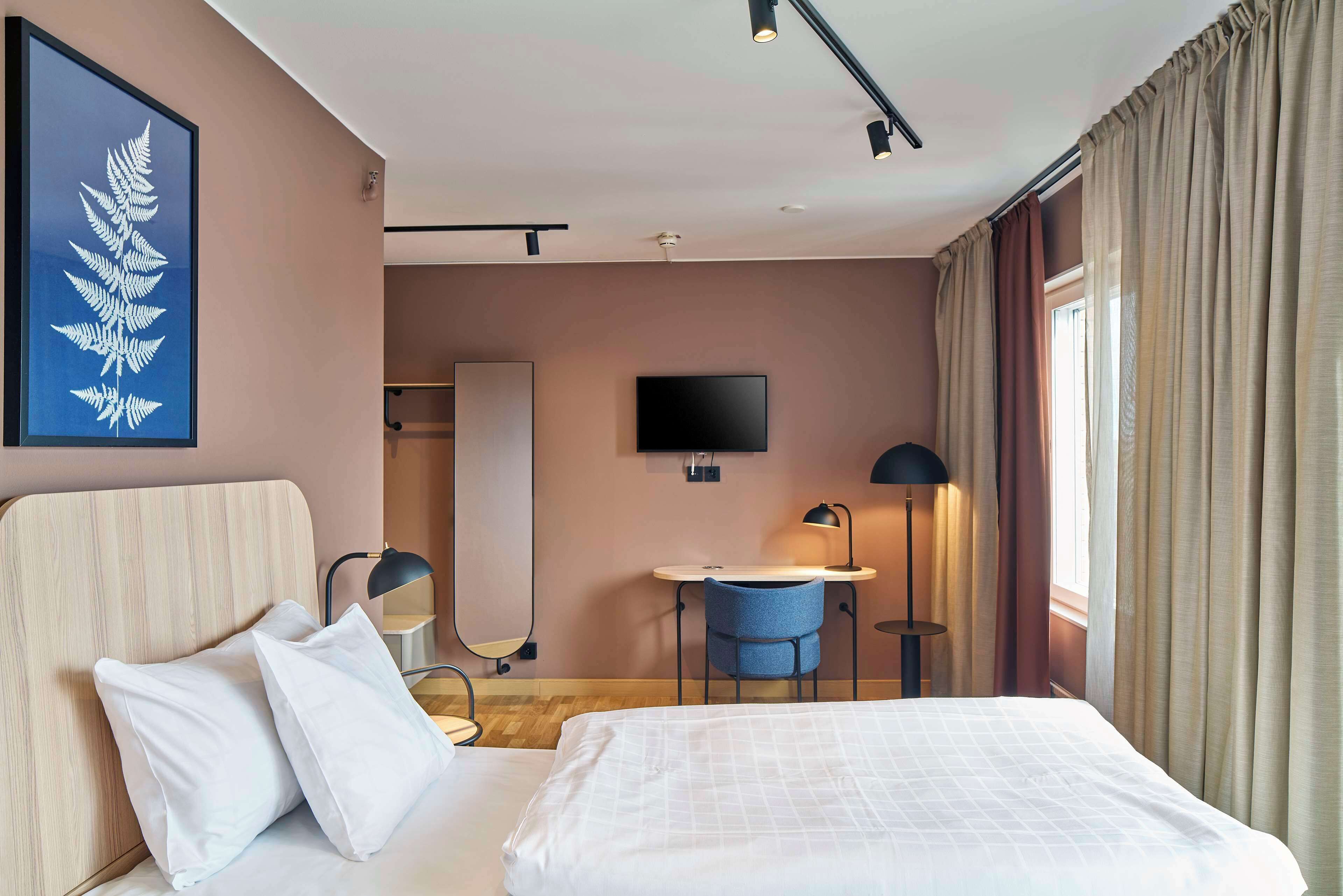 Ett hotellrum med en säng, ett skrivbord, en stol, en tv och en inramad växt på väggen.