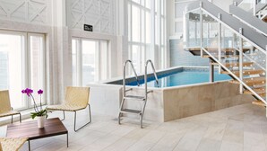 Un área moderna de piscina interior con una escalera de cristal, sillas de descanso y vistas a la ciudad.
