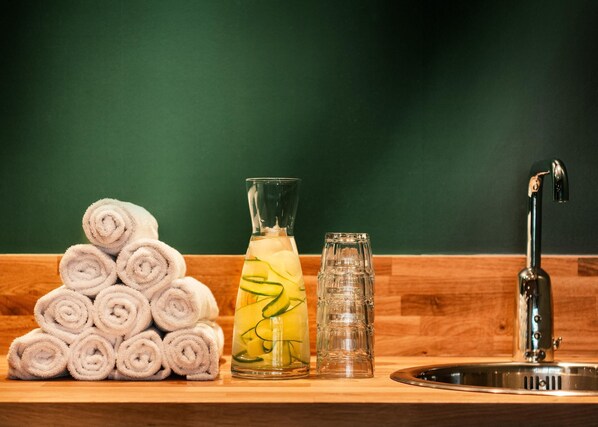 Una pila de toallas enrolladas, una botella de vidrio con líquido amarillo y una jarra de vidrio sobre una encimera de madera con un lavabo.