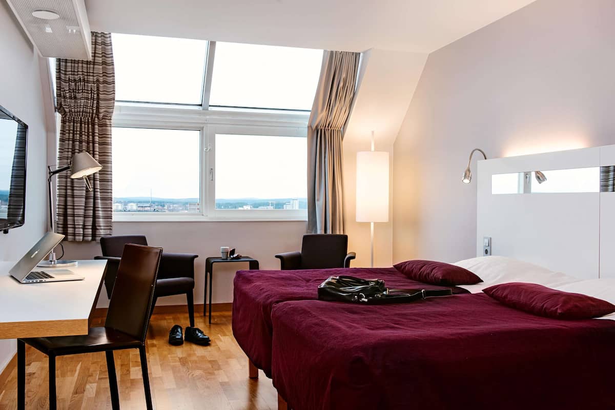 A hotel room with a bed, desk, chair, and a view of the city through the window.