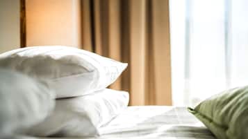Una cama bien hecha con almohadas blancas y verdes, una lámpara y una ventana con cortinas al fondo.