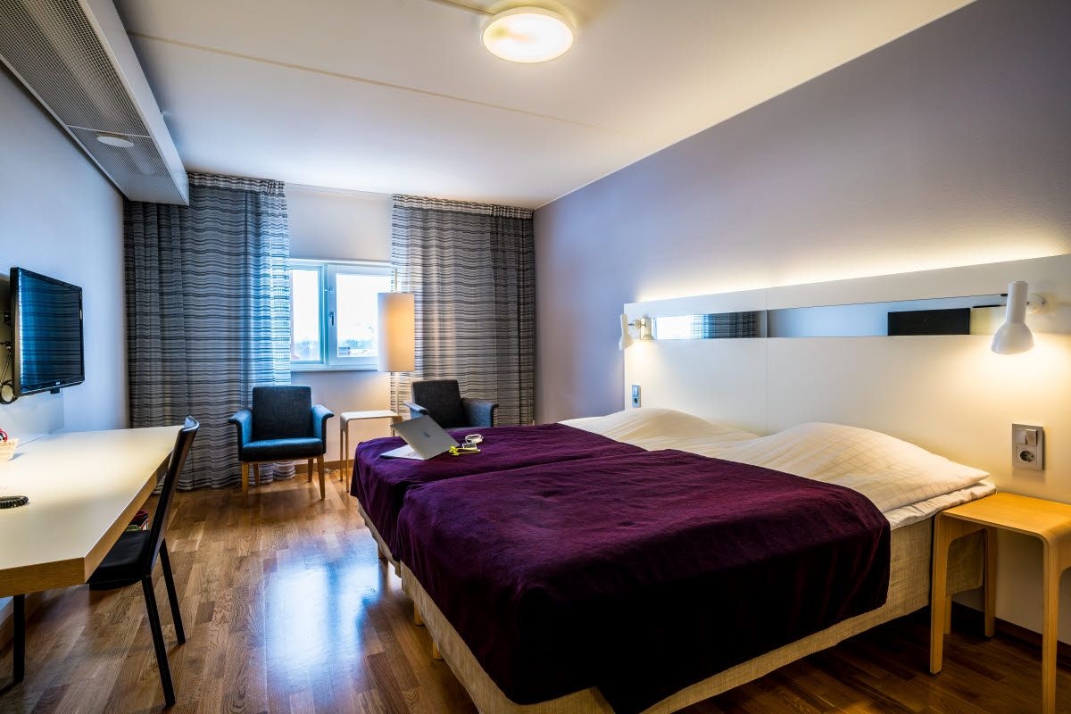 Moderni hotellihuone, jossa on sänky, työpöytä, tuoli ja ikkuna, jossa on verhot.