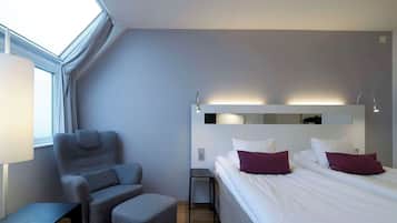 A modern hotel room with a skylight, a grey armchair, a small table, a bed with white and purple pillows, and a nightstand with a lamp.