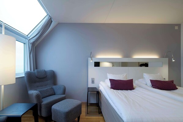 A modern hotel room with a skylight, a grey armchair, a small table, a bed with white and purple pillows, and a nightstand with a lamp.