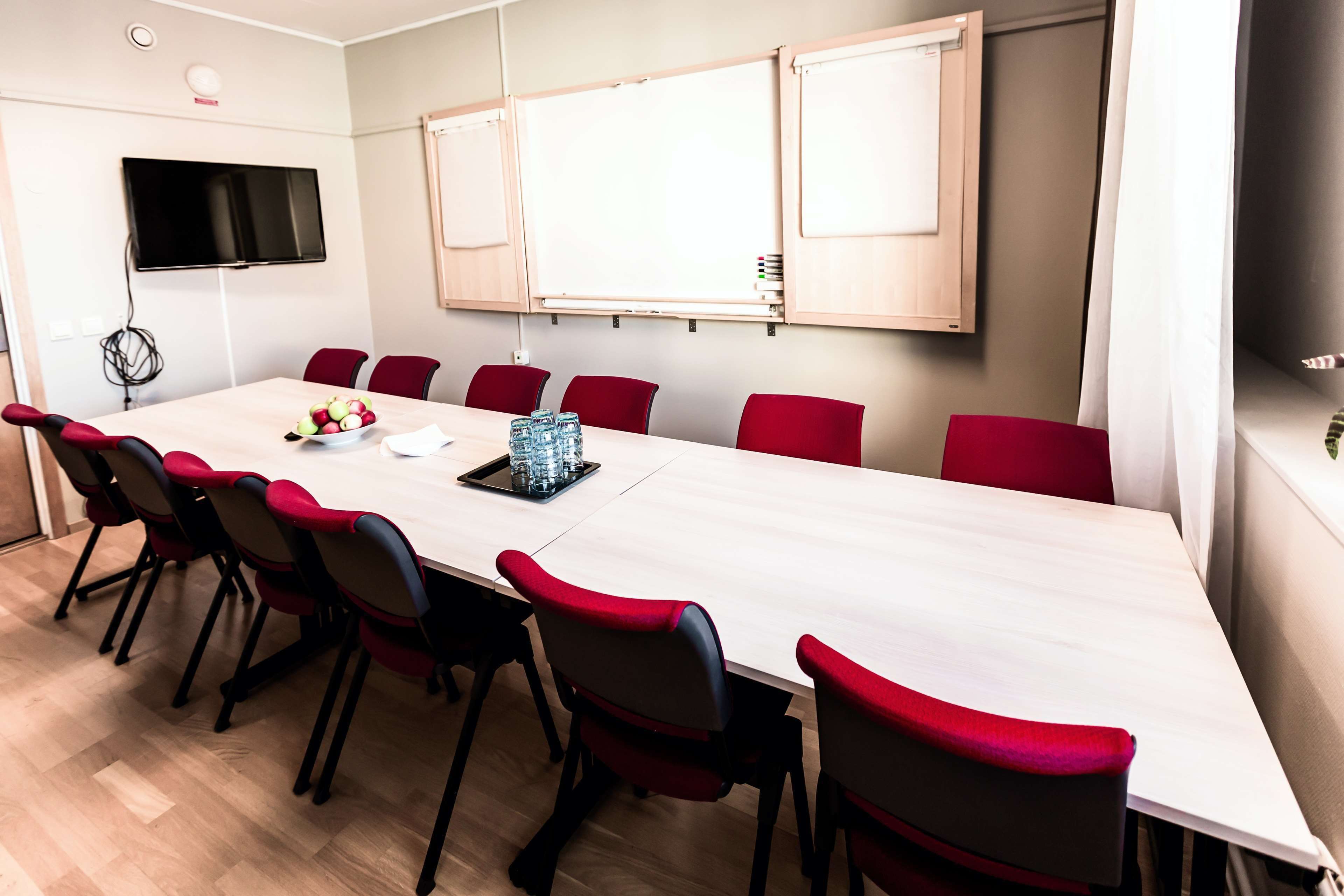 Un salón de conferencias con una larga mesa, sillas rojas, una pizarra y un televisor montado en la pared.
