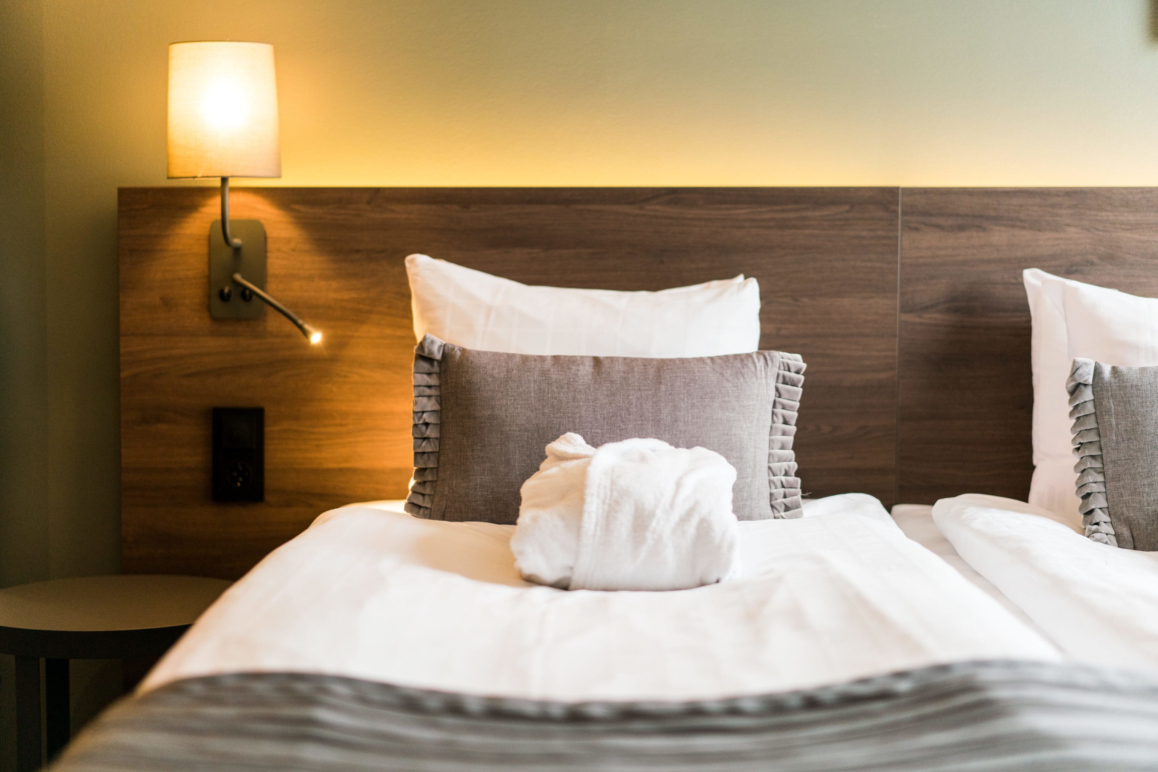 A hotel room with a wooden headboard, a bedside lamp, and a neatly made bed with pillows and a towel.