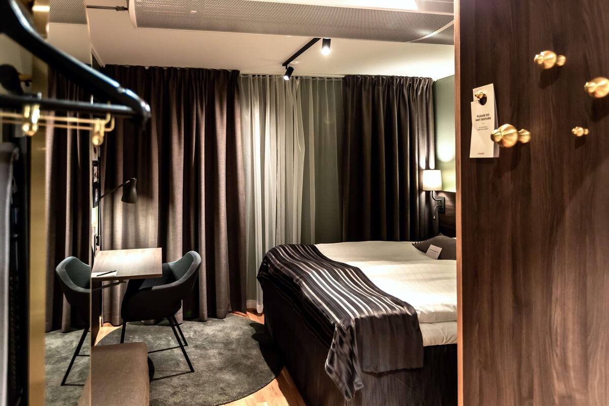 A hotel room with a bed, a desk, two chairs, a desk lamp, a coat rack, and a door with a sign.
