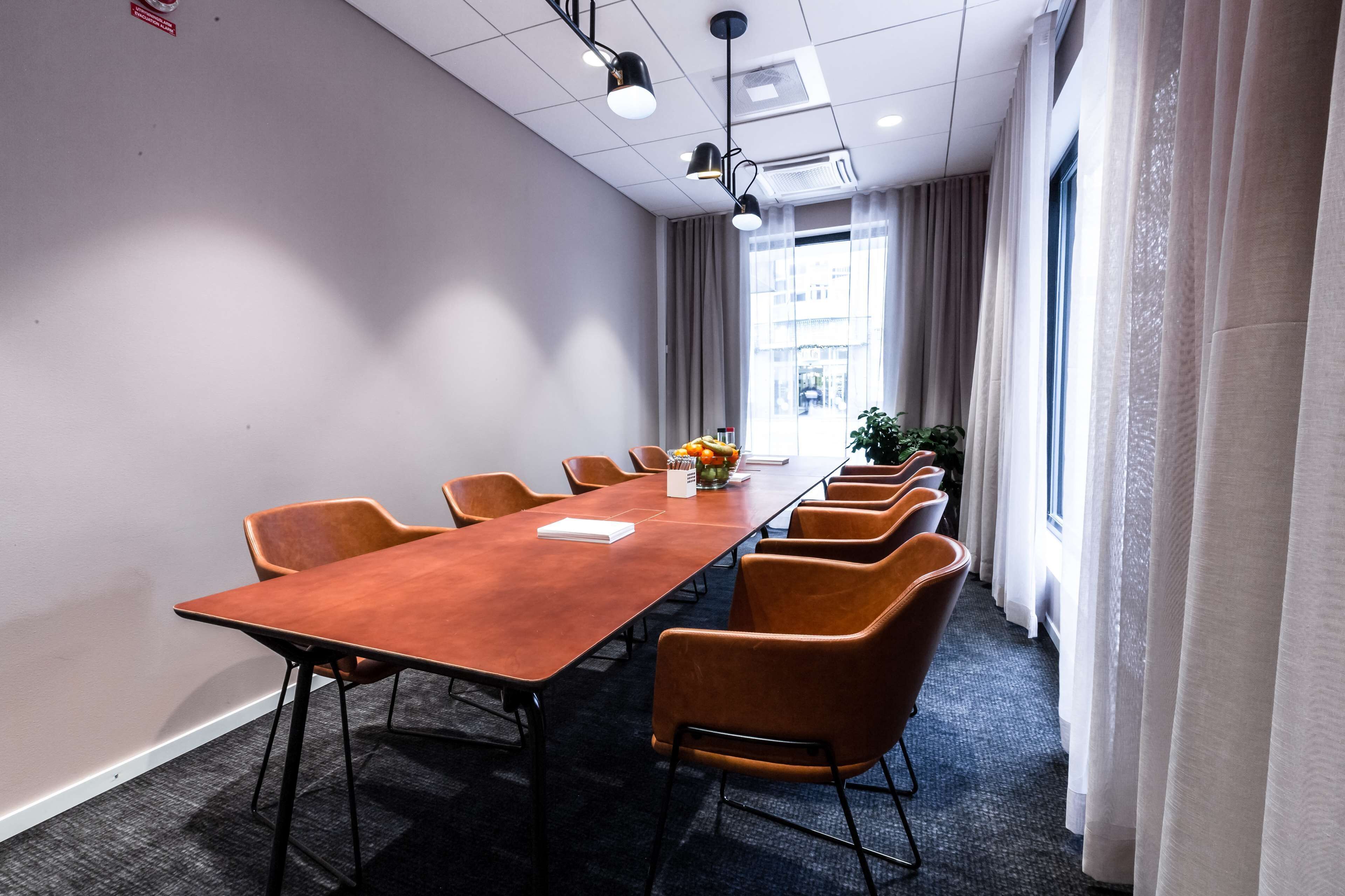 A conference room with a long table and several chairs, a plant, and a window with curtains.