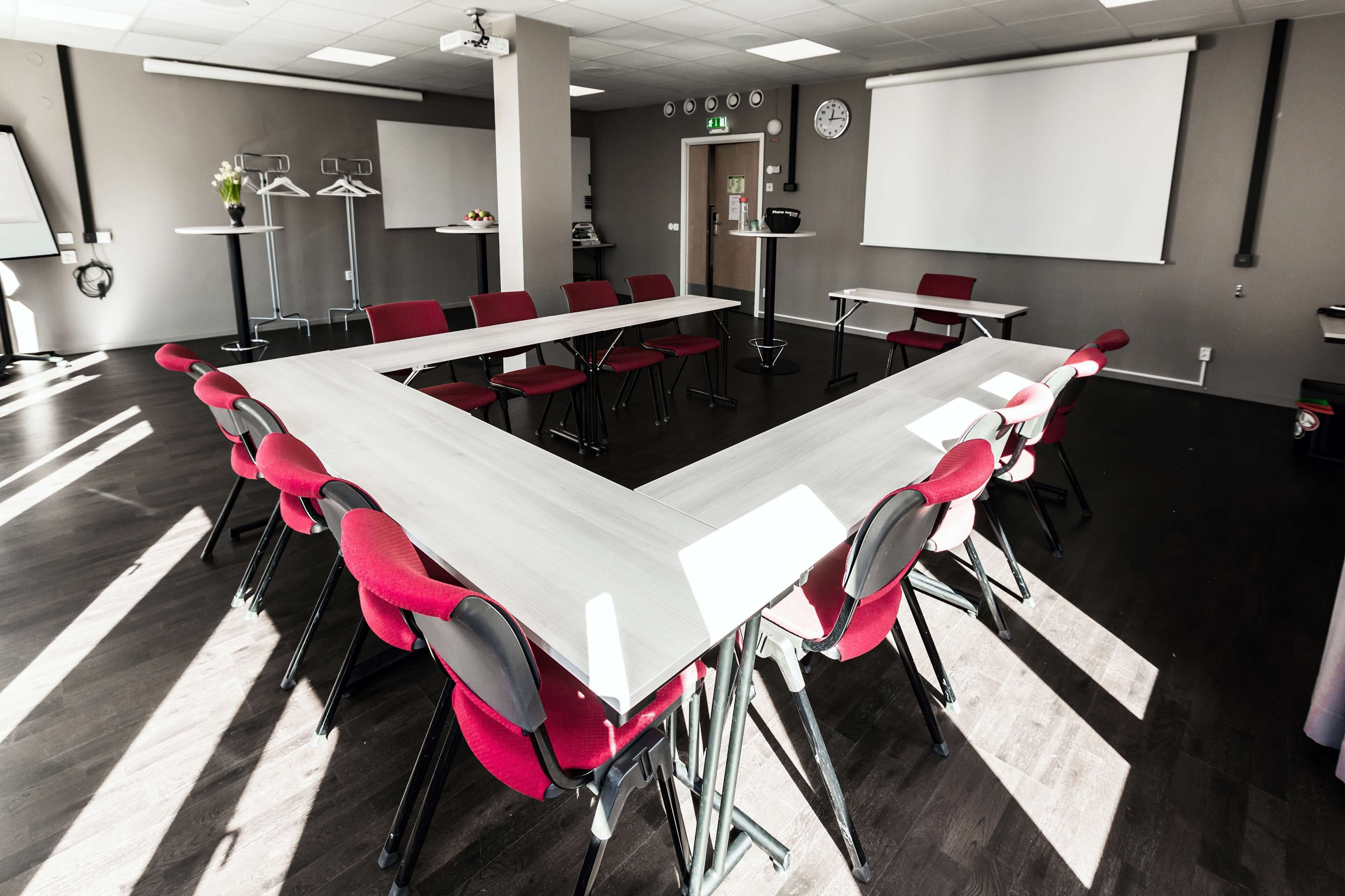 Un salón de conferencias con una larga mesa, sillas rojas, una pantalla de proyector y una pizarra.