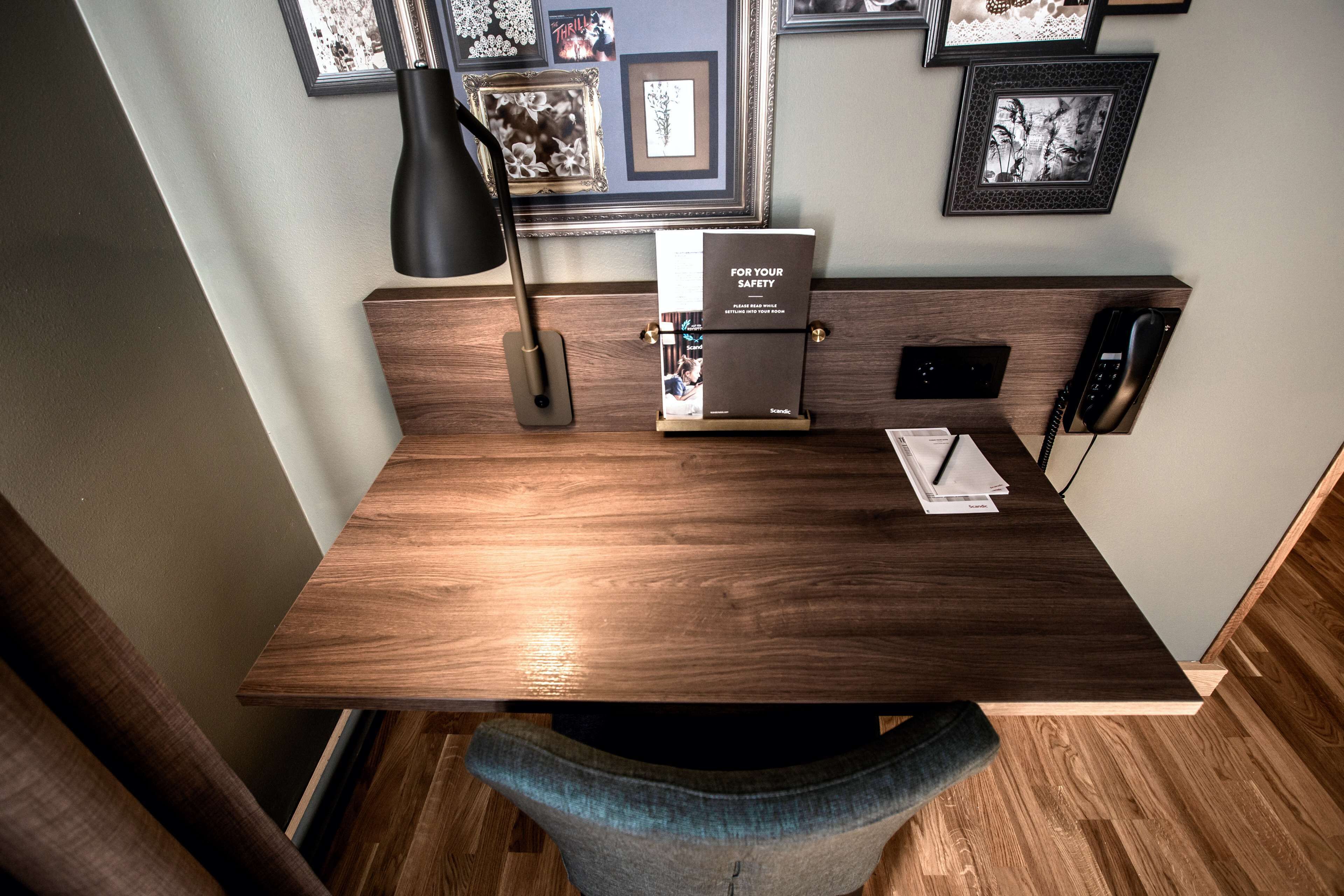 A wooden desk with a black lamp, a phone, and a framed book titled "FOR YOUR SAFETY" on the wall behind it.